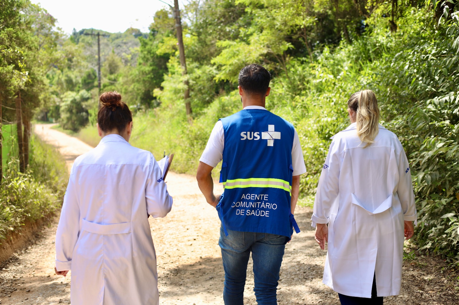 A imagem mostra três profissionais de saúde caminhando por uma estrada de terra em uma área rural, cercada por vegetação densa e verde.  Eles são vistos de costas. Ao centro, um homem veste um colete azul com a identificação do SUS e a inscrição “Agente Comunitário de Saúde”. Ao seu lado, duas mulheres usam jalecos brancos; uma delas segura uma prancheta, sugerindo que estão em atividade de campo.  O cenário é ensolarado e transmite a ideia de deslocamento no território, provavelmente para visitas domiciliares ou acompanhamento de moradores. A cena reforça o trabalho da atenção básica e da estratégia de saúde da família, especialmente em áreas mais afastadas ou de difícil acesso.