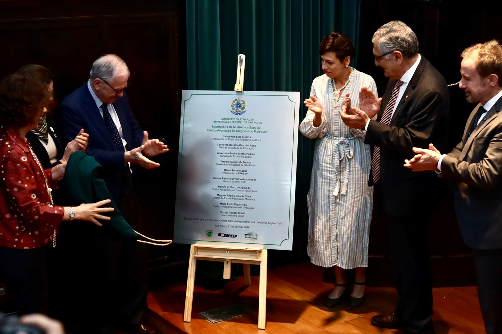 A imagem mostra uma cerimônia formal de inauguração ou homenagem em ambiente institucional. No centro, há uma placa comemorativa apoiada em um cavalete de madeira, com o brasão oficial do Brasil no topo e um texto detalhando a ocasião — possivelmente a criação ou entrega de um equipamento ou espaço ligado à educação ou à saúde.  Ao redor da placa, seis pessoas bem vestidas participam do momento. À esquerda, um homem idoso de terno parece ter acabado de puxar uma cortina verde que revelava a placa, enquanto os demais — homens e mulheres em trajes sociais — aplaudem o ato. A cena transmite solenidade e celebração.  O ambiente é interno, com piso de madeira e cortinas verdes ao fundo, sugerindo um auditório ou sala de eventos oficial. A iluminação destaca a placa e os participantes, reforçando o caráter simbólico e institucional da cerimônia.
