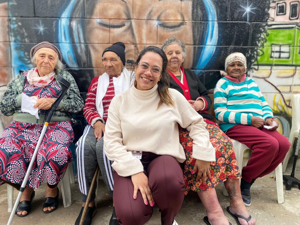 A médica da família Sabrina Teixeira aparece agachada ao centro da imagem, sorrindo ao lado de quatro idosas acompanhadas pela equipe de saúde. Ao fundo, um mural colorido com o rosto de uma mulher de olhos fechados reforça a atmosfera de acolhimento e cuidado comunitário. As pacientes estão sentadas em cadeiras na área externa da unidade, em um momento que transmite vínculo, proximidade e convivência no território.