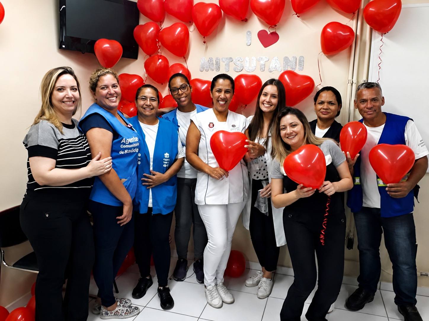 A foto mostra um grupo de nove pessoas reunidas em um ambiente interno da Unidade Básica de Saúde (UBS) Mitsutani. O local está decorado para uma celebração temática. Na parede ao fundo há muitos balões vermelhos em formato de coração, além de um cartaz com o nome “Mitsutani”, sugerindo uma homenagem ou evento especial. A decoração cria um clima festivo e acolhedor. O grupo está posicionado lado a lado, sorrindo para a câmera. Algumas pessoas usam coletes azuis com identificação de agente comunitário de saúde, enquanto outras vestem roupas casuais ou uniformes brancos de profissionais da saúde. Várias seguram balões vermelhos em formato de coração nas mãos. No centro, uma mulher de uniforme branco se destaca, cercada pelos colegas, reforçando a sensação de celebração em equipe. O ambiente transmite união, confraternização e valorização do trabalho coletivo.