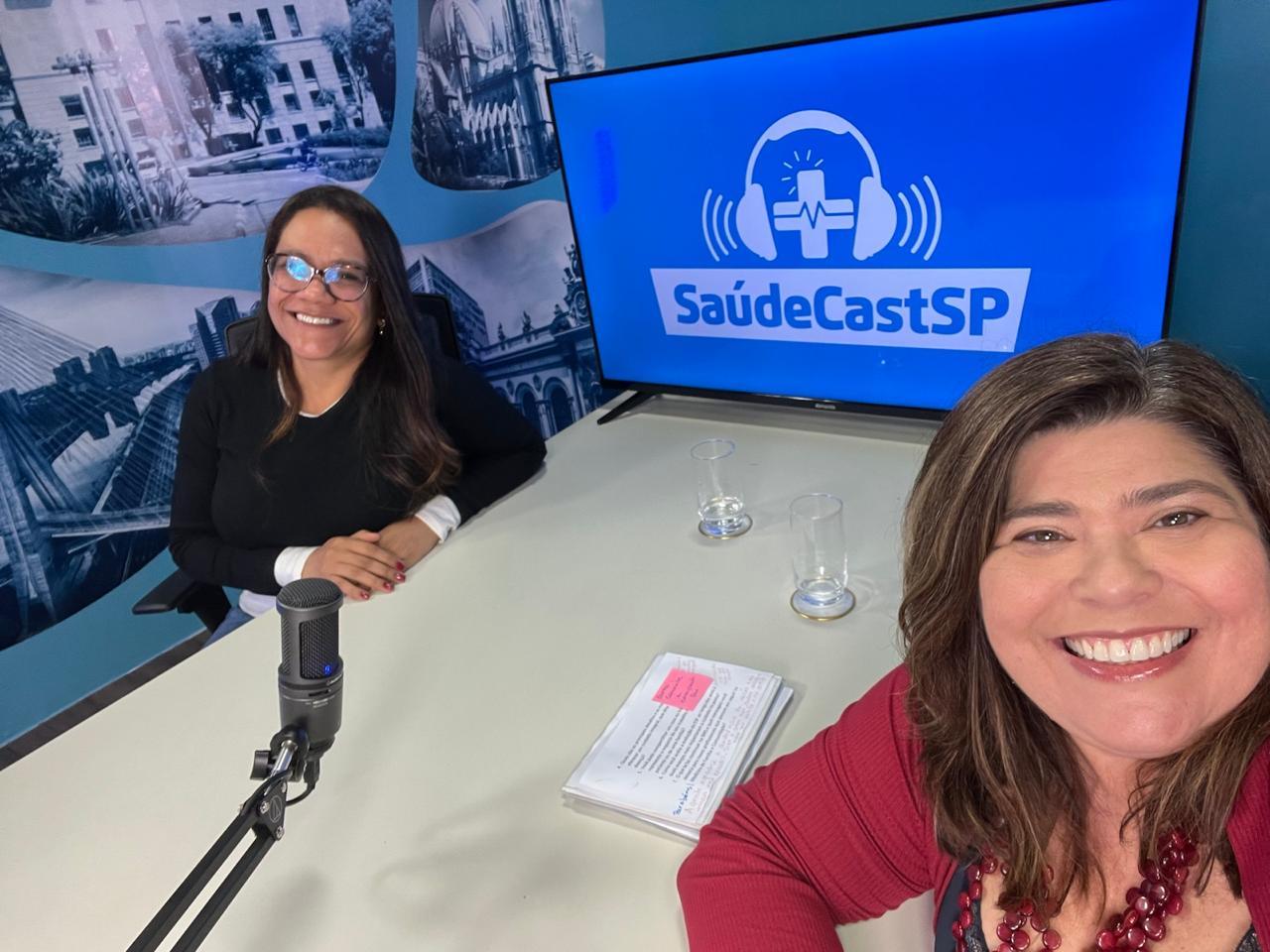 A imagem mostra duas mulheres sorrindo em um ambiente que parece ser um estúdio de gravação de podcast ou conteúdo em áudio. Ambas estão sentadas em torno de uma mesa, onde há um microfone de gravação, dois copos de água e anotações impressas. Ao fundo, um monitor exibe a identidade visual “SaúdeCastSP”, acompanhada de um ícone de fones de ouvido com um símbolo de saúde ao centro, sugerindo que o espaço é voltado à produção de conteúdo sobre saúde. A parede também possui imagens em tons de azul que remetem à cidade de São Paulo e a cenários urbanos. A cena transmite um clima de conversa, entrevista ou gravação de programa institucional, com um ambiente acolhedor e profissional voltado à comunicação em saúde.