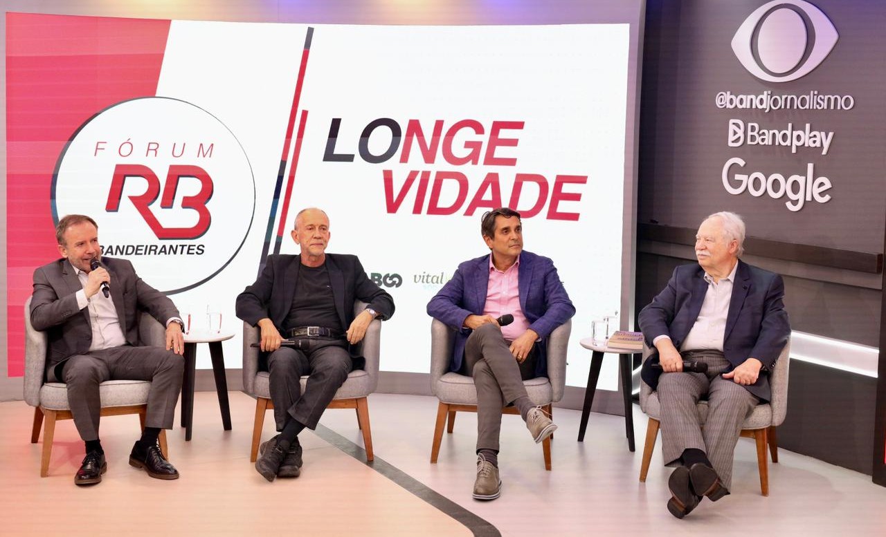 A imagem mostra um painel de debates realizado em um estúdio ou auditório de comunicação, com quatro homens sentados em poltronas no palco, participando de uma conversa mediada. Um deles segura um microfone e parece estar falando, enquanto os demais acompanham a discussão.  Ao fundo, há um telão com os dizeres “Fórum RB Bandeirantes” e o tema “Longe Vida”, sugerindo um evento voltado a saúde, qualidade de vida, envelhecimento ou longevidade. Também aparecem logotipos ligados ao grupo de comunicação, como Band Jornalismo, Bandplay e Google.  O ambiente tem aparência de evento institucional ou fórum temático, com iluminação profissional, mesas laterais com copos de água e uma composição visual típica de transmissões, entrevistas ou debates públicos.