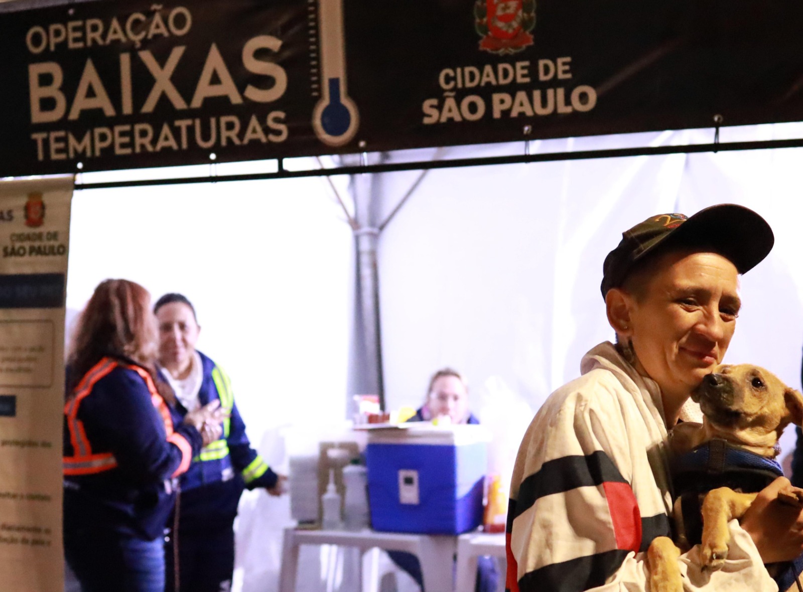 A imagem mostra uma ação da “Operação Baixas Temperaturas”, iniciativa da Cidade de São Paulo voltada ao acolhimento e proteção de pessoas em períodos de frio intenso. Na parte superior, há uma faixa com os dizeres “Operação Baixas Temperaturas” e o brasão da cidade.  Em primeiro plano, aparece uma pessoa usando boné e casaco claro, segurando um cachorro no colo, em um momento que transmite acolhimento e cuidado. Ao fundo, dentro de uma estrutura de atendimento montada provisoriamente, há profissionais conversando e uma mesa com materiais de apoio, sugerindo um ponto de assistência.  O cenário remete a uma ação social ou humanitária voltada ao atendimento de populações em situação de vulnerabilidade durante dias de temperaturas mais baixas, incluindo atenção também aos animais de estimação que acompanham seus tutores.