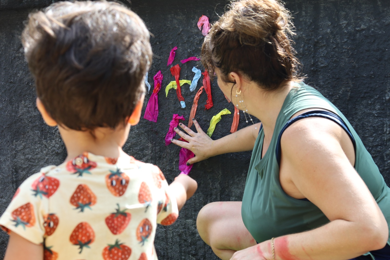 A imagem mostra uma mulher e uma criança pequena interagindo com uma superfície escura, que parece um muro ou painel, onde há pedaços coloridos de papel ou material decorativo fixados, formando uma composição artística ou atividade lúdica.  A mulher está agachada, com uma das mãos apoiada no painel, observando a atividade, enquanto a criança, em primeiro plano e usando uma camiseta com estampa de morangos, aponta ou participa da montagem. Ambos parecem envolvidos em uma ação de caráter educativo, criativo ou recreativo ao ar livre.  A cena transmite um clima de interação, aprendizado e construção conjunta, com foco na experiência compartilhada entre adulto e criança.