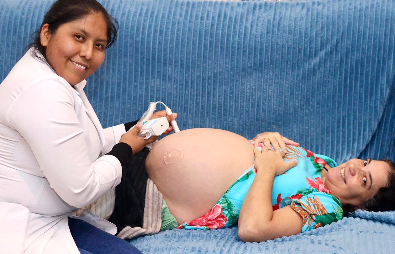 A imagem mostra uma gestante deitada confortavelmente em uma maca ou poltrona reclinável, sorrindo para a câmera, enquanto uma profissional de saúde realiza um exame em sua barriga com um aparelho portátil, possivelmente relacionado ao acompanhamento pré-natal.  A profissional, vestindo jaleco branco, também olha para a câmera e segura um equipamento com um sensor apoiado sobre o abdômen da paciente, onde é possível ver o gel utilizado no procedimento. A gestante usa um vestido estampado colorido e mantém as mãos apoiadas sobre a região do tórax.  O ambiente transmite uma cena de cuidado e acompanhamento em saúde materna, sugerindo um momento de atendimento pré-natal, monitoramento do bebê ou avaliação da gestação em um serviço de saúde.