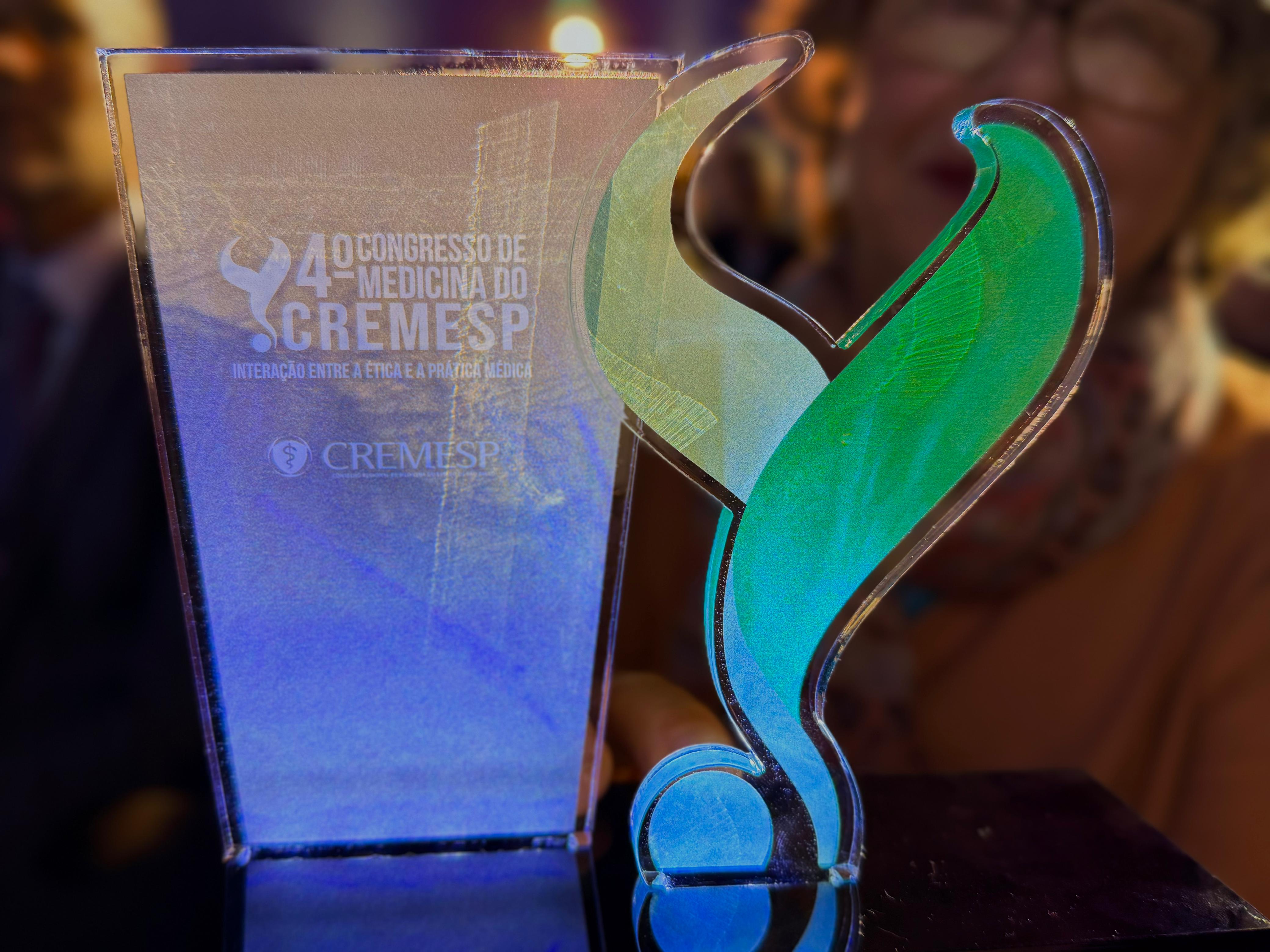 Troféu em acrílico transparente com detalhes em verde e azul, iluminado por luzes quentes ao fundo. Na peça está gravada a inscrição “4º Congresso de Medicina do Cremesp – Interação entre a Ética e a Prática Médica”, além da logomarca do Cremesp. A fotografia destaca o reconhecimento institucional ligado ao evento voltado à discussão de temas da medicina, ética e inovação em saúde