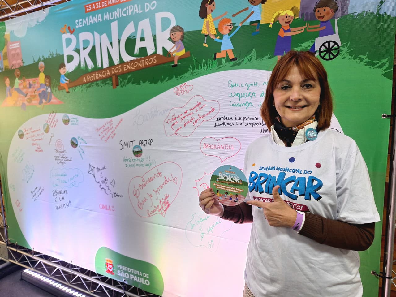 A imagem mostra a participação da Secretaria Municipal da Saúde (SMS) em atividade da Semana Municipal do Brincar, realizada em alusão ao Dia Mundial do Brincar. Em primeiro plano, a diretora da Divisão de Saúde por Ciclos de Vida e responsável pela Área Técnica de Saúde da Criança e do Adolescente da SMS, Athenê Maria de Marco Mauro, posa diante de um painel temático do evento, segurando material da campanha. Ao fundo, o painel colorido destaca o tema “A Potência dos Encontros” e reúne mensagens escritas pelos participantes sobre a importância do brincar para o desenvolvimento infantil, a convivência e a promoção da saúde.