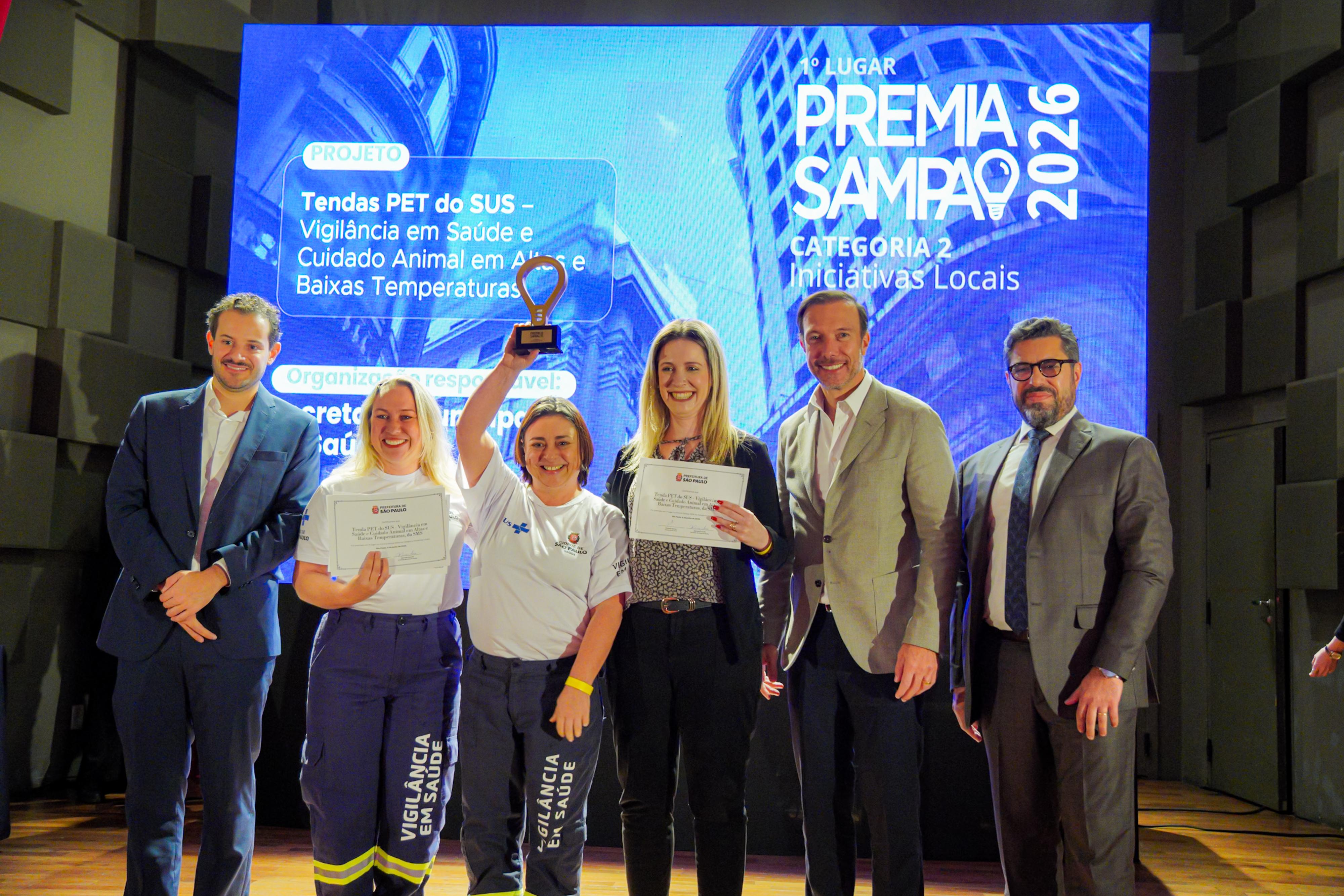 A imagem mostra uma cerimônia de premiação realizada em um auditório, com seis pessoas posando no palco diante de um telão que exibe o resultado do Premia Sampa 2026. Ao centro, integrantes da equipe vencedora seguram certificados e um troféu erguido acima da cabeça, celebrando a conquista.  No telão ao fundo, é possível ler que o projeto premiado foi “Tendas PET do SUS – Vigilância em Saúde e Cuidado Animal em Ações de Baixas Temperaturas”, vencedor do 1º lugar na Categoria 2 – Iniciativas Locais. A tela também indica a vinculação do projeto à Secretaria Municipal da Saúde de São Paulo.  As duas profissionais ao centro vestem camisetas da Vigilância em Saúde e exibem os certificados da premiação, enquanto os demais participantes, trajando roupas sociais, acompanham o momento de reconhecimento institucional. A iluminação do palco e a projeção ao fundo reforçam o caráter formal do evento.