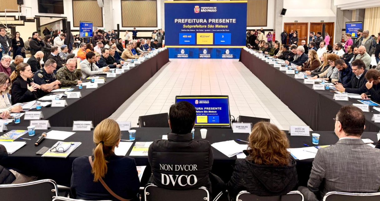 A imagem mostra uma grande reunião institucional realizada em um auditório ou centro de convenções. Os participantes estão distribuídos ao redor de mesas dispostas em formato de