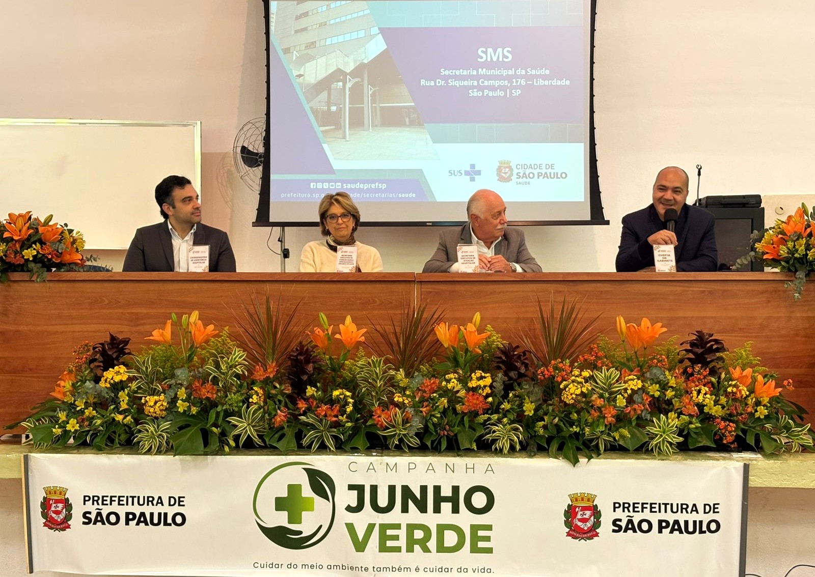 A imagem mostra uma mesa de autoridades durante um evento institucional realizado em auditório ou salão de conferências. Quatro pessoas estão sentadas atrás de uma longa bancada de madeira, ornamentada com um grande arranjo de flores e folhagens em tons de laranja, amarelo e verde. Ao fundo, um telão exibe uma apresentação da Secretaria Municipal da Saúde (SMS) de São Paulo, com referência ao endereço da sede da pasta, na Rua Dr. Siqueira Campos, na Liberdade. Também aparecem os logotipos do SUS e da Prefeitura de São Paulo. Na parte frontal da mesa há uma faixa da campanha “Junho Verde”, acompanhada do slogan “Cuidar do meio ambiente também é cuidar da vida”, além dos brasões da Prefeitura de São Paulo. O homem à direita está com um microfone em mãos e parece estar discursando ou participando de uma mesa-redonda, enquanto os demais acompanham a atividade.