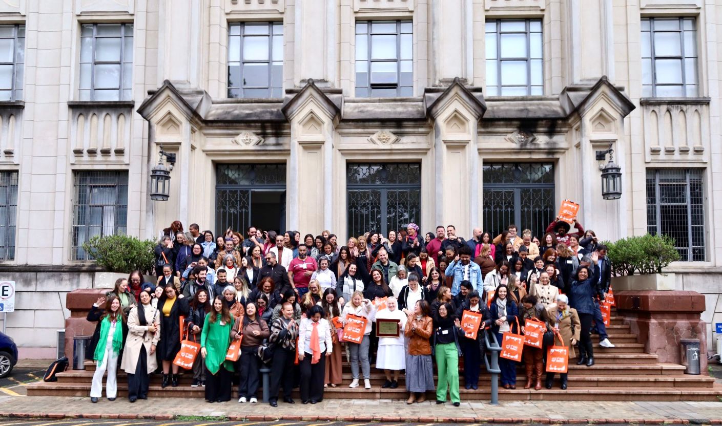 A imagem mostra um grande grupo de pessoas reunidas para uma foto oficial em frente à entrada de um edifício histórico de grande porte, com fachada em estilo clássico, marcada por colunas, janelas altas e detalhes ornamentais. Os participantes estão distribuídos pelos degraus da escadaria principal. A maioria sorri, conversa ou acena para a câmera, criando um clima descontraído e de confraternização. Algumas pessoas seguram sacolas laranja personalizadas, sugerindo que participaram de um evento, congresso ou encontro institucional. No centro da primeira fileira, uma pessoa veste uma roupa branca e segura uma placa comemorativa, em torno da qual os demais se concentram. O grupo é bastante diverso, formado por homens e mulheres de diferentes idades e etnias. Muitos utilizam crachás de identificação, reforçando o caráter organizado do evento. As vestimentas variam entre trajes sociais, casuais e esportivos, com predominância de casacos, compatíveis com um dia de temperatura amena.