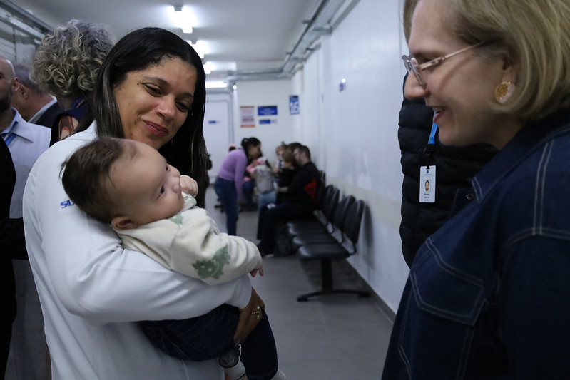 A imagem mostra um momento de interação em uma unidade de saúde. Em primeiro plano, uma profissional de saúde, vestindo jaleco branco, segura um bebê no colo enquanto sorri para uma mulher posicionada à direita da foto. A mulher, que usa óculos e um casaco azul-marinho, também sorri e observa a criança, transmitindo um clima de acolhimento e proximidade. O bebê, vestido com um casaco claro e calça escura, está voltado para a mulher, parecendo atento à interação. Ao fundo, há um corredor com cadeiras de espera ocupadas por outras pessoas, além de profissionais e acompanhantes circulando pelo local, o que sugere o funcionamento regular da unidade. A iluminação é feita por lâmpadas fluorescentes no teto, e o ambiente tem paredes brancas e piso cinza, típicos de um equipamento público de saúde.