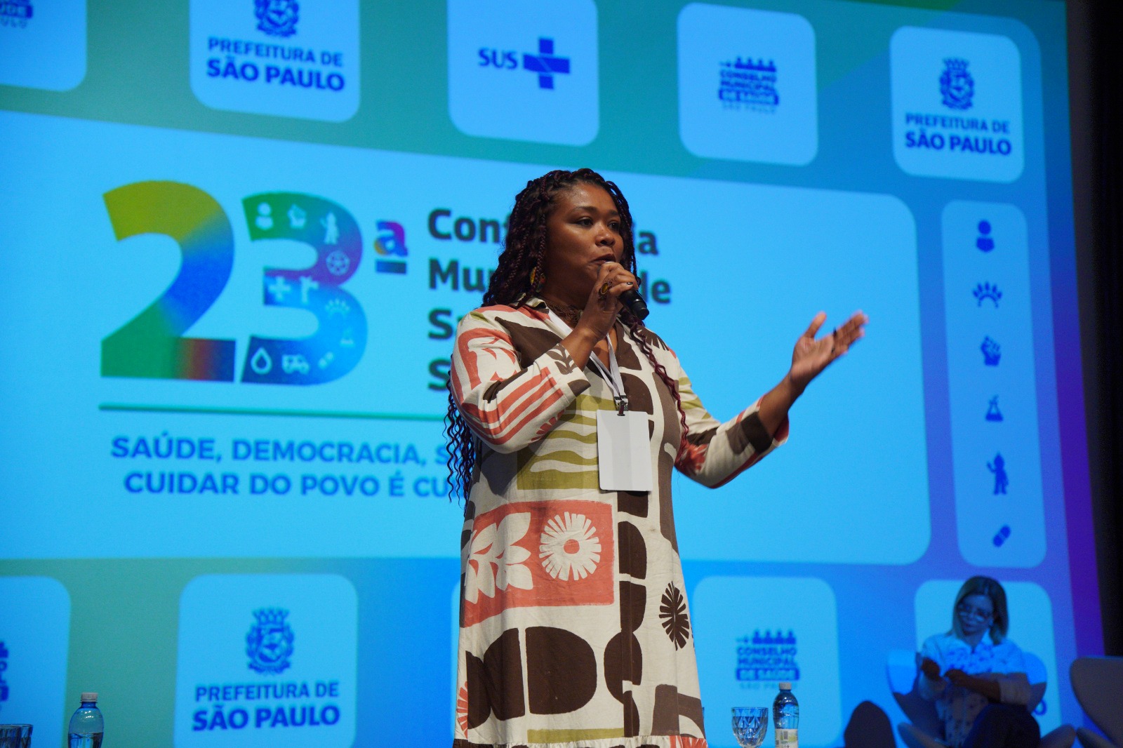 A imagem mostra uma mulher em pé sobre um palco, falando ao microfone durante um evento da área da saúde. Ela veste um vestido estampado em tons de branco, marrom, verde e terracota, usa um crachá de identificação pendurado no pescoço e faz um gesto com a mão esquerda enquanto se dirige ao público, transmitindo uma postura segura e participativa.  Ao fundo, um grande telão exibe a identidade visual da 23ª Conferência Municipal de Saúde de São Paulo, com destaque para o número 
