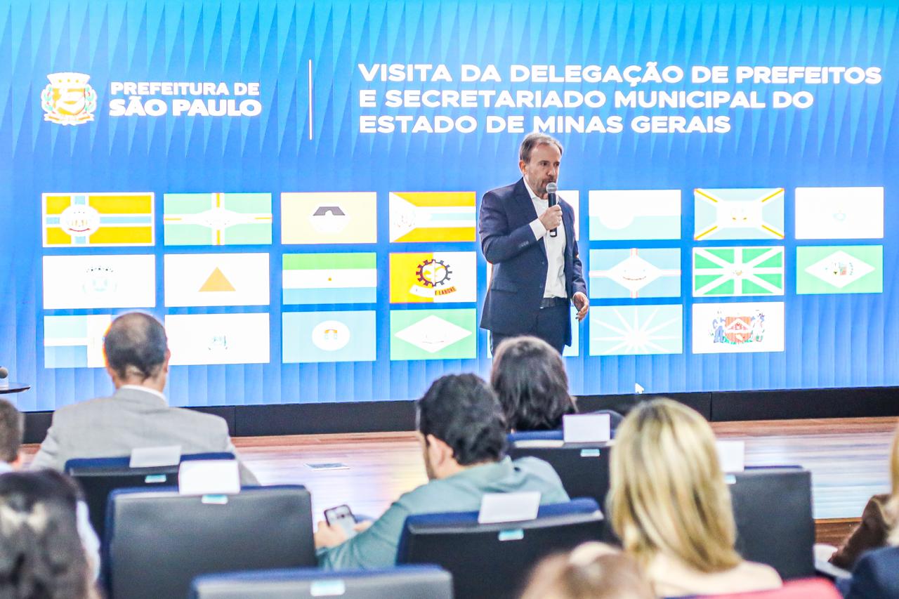 A imagem mostra um auditório durante uma apresentação institucional da Prefeitura de São Paulo. Em primeiro plano, aparecem participantes sentados, voltados para o palco, acompanhando a exposição.  No centro da cena, um homem de terno segura um microfone e faz uma apresentação diante de um grande painel de LED. Ao fundo, o telão exibe a identidade visual da Prefeitura de São Paulo e a mensagem: 