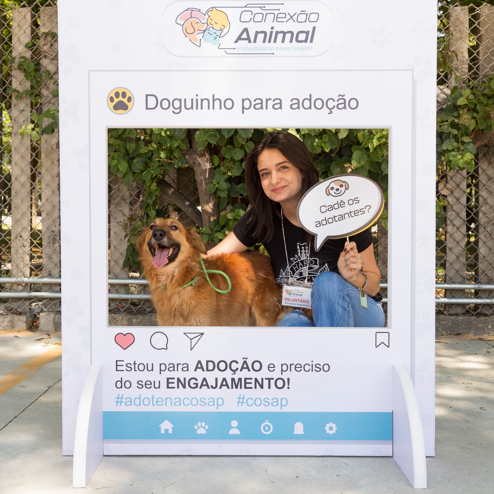 A foto mostra uma ação de incentivo à adoção de cães em um evento ao ar livre. Em destaque, há um painel cenográfico em formato de publicação de rede social, com a identidade visual do projeto Conexão Animal e a frase 