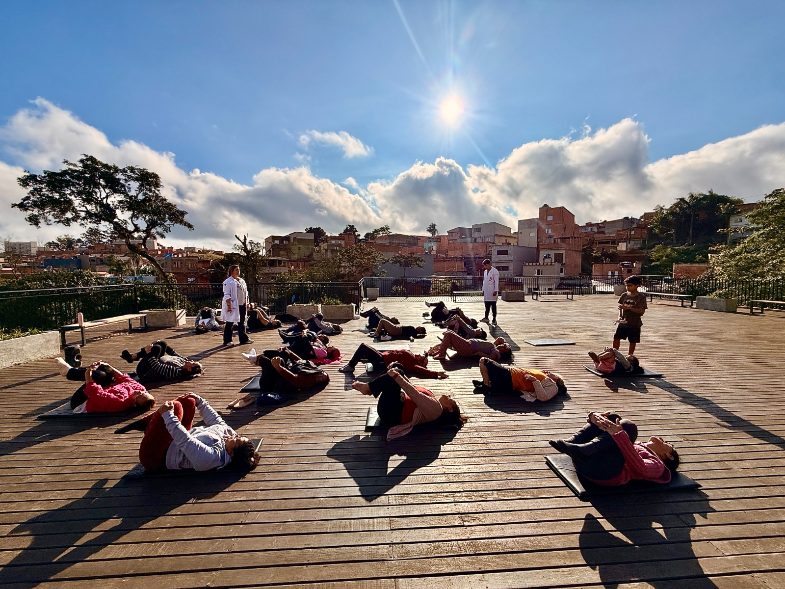 A imagem mostra uma atividade coletiva de exercícios ou alongamento realizada ao ar livre em um amplo deck de madeira.  No primeiro plano, diversas pessoas estão deitadas sobre colchonetes, realizando um exercício em que abraçam os joelhos junto ao peito. As pessoas estão distribuídas pelo espaço, mantendo certa distância entre si. As longas sombras projetadas no piso indicam que o sol está relativamente baixo no céu, sugerindo o início da manhã ou o fim da tarde.  Ao centro da cena, duas pessoas vestindo jalecos brancos parecem acompanhar ou orientar a atividade. À direita, uma criança está em pé observando o grupo, enquanto outra pessoa também permanece em pé próxima ao centro.  O ambiente é aberto, cercado por um guarda-corpo metálico, com bancos e grandes vasos ao redor. Ao fundo, há um bairro com casas e edifícios de diferentes tamanhos distribuídos sobre um terreno em aclive. À esquerda, destaca-se uma árvore de copa ampla.  O céu ocupa boa parte da imagem e chama a atenção pelo azul intenso, com nuvens volumosas iluminadas pelo sol, que aparece próximo ao topo da fotografia, criando um forte brilho e raios de luz. A combinação da iluminação, das sombras e da paisagem transmite a sensação de uma atividade ao ar livre em um dia claro e agradável.