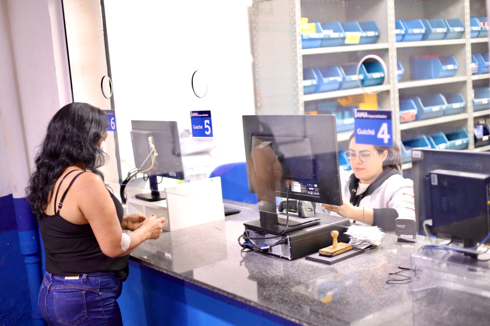 Mulher é atendida no guichê de uma farmácia da rede municipal de saúde. Do outro lado do balcão, uma farmacêutica realiza a dispensação de medicamentos por meio do sistema informatizado. Ao fundo, prateleiras organizadas com caixas e compartimentos para armazenamento de medicamentos evidenciam a estrutura do serviço farmacêutico da rede.