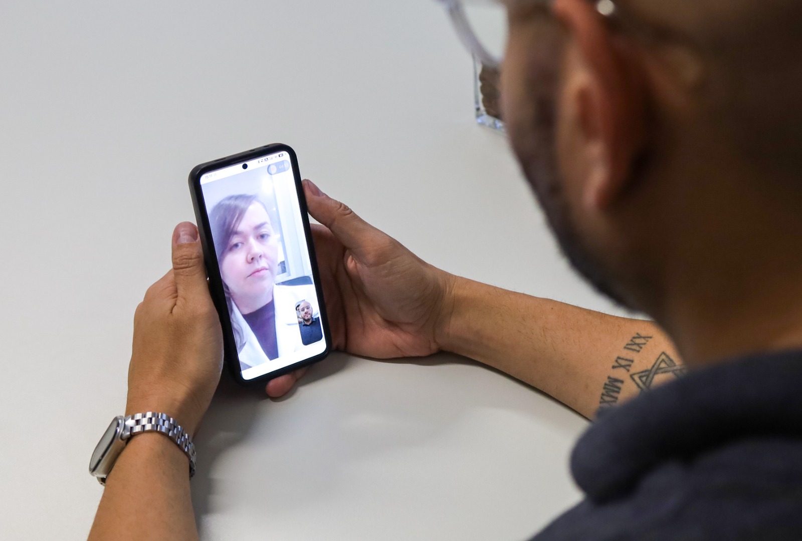 A imagem mostra uma pessoa sentada à mesa, vista de costas e de lado, segurando um smartphone com as duas mãos. Na tela do aparelho aparece uma videochamada, com uma mulher ocupando a maior parte da imagem e, no canto inferior, a pequena janela de vídeo do outro participante.  A cena sugere um atendimento ou consulta por telemedicina, destacando o uso da tecnologia para possibilitar uma conversa à distância. A composição é simples, com fundo claro e iluminação suave, e coloca o celular e a videochamada como elementos centrais da fotografia.
