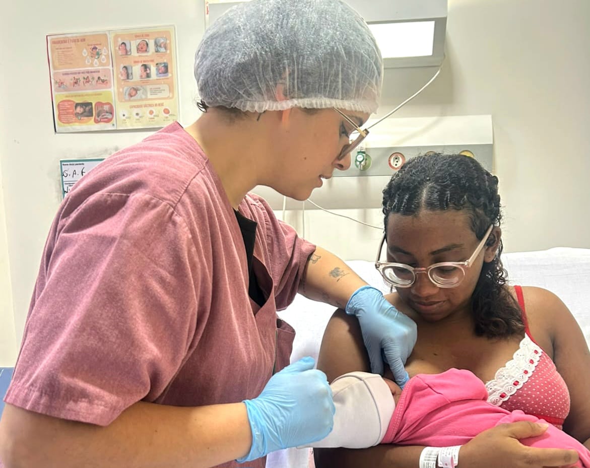 Em ambiente hospitalar, uma profissional de saúde usando touca, óculos, avental rosa e luvas azuis orienta uma mulher durante a amamentação. A mãe, sentada em uma cama, segura o bebê no colo, coberto por uma manta rosa, enquanto recebe auxílio para posicioná-lo junto ao peito. A cena representa o cuidado e o apoio à amamentação oferecidos por profissionais de saúde.