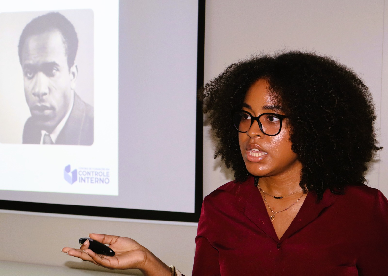 Uma mulher negra, de cabelos crespos volumosos, usa óculos de armação preta e uma camisa bordô enquanto faz uma apresentação. Ela está voltada ligeiramente para a esquerda, com a boca entreaberta, como se estivesse falando, e segura um pequeno controle de apresentação na mão direita, com o braço estendido à frente. Usa colares delicados e pulseiras. Ao fundo, uma tela de projeção exibe a fotografia em preto e branco de um homem e o logotipo do Departamento de Controle Interno. A cena ocorre em um ambiente fechado, com iluminação que destaca a palestrante e transmite a impressão de uma exposição ou palestra profissional.