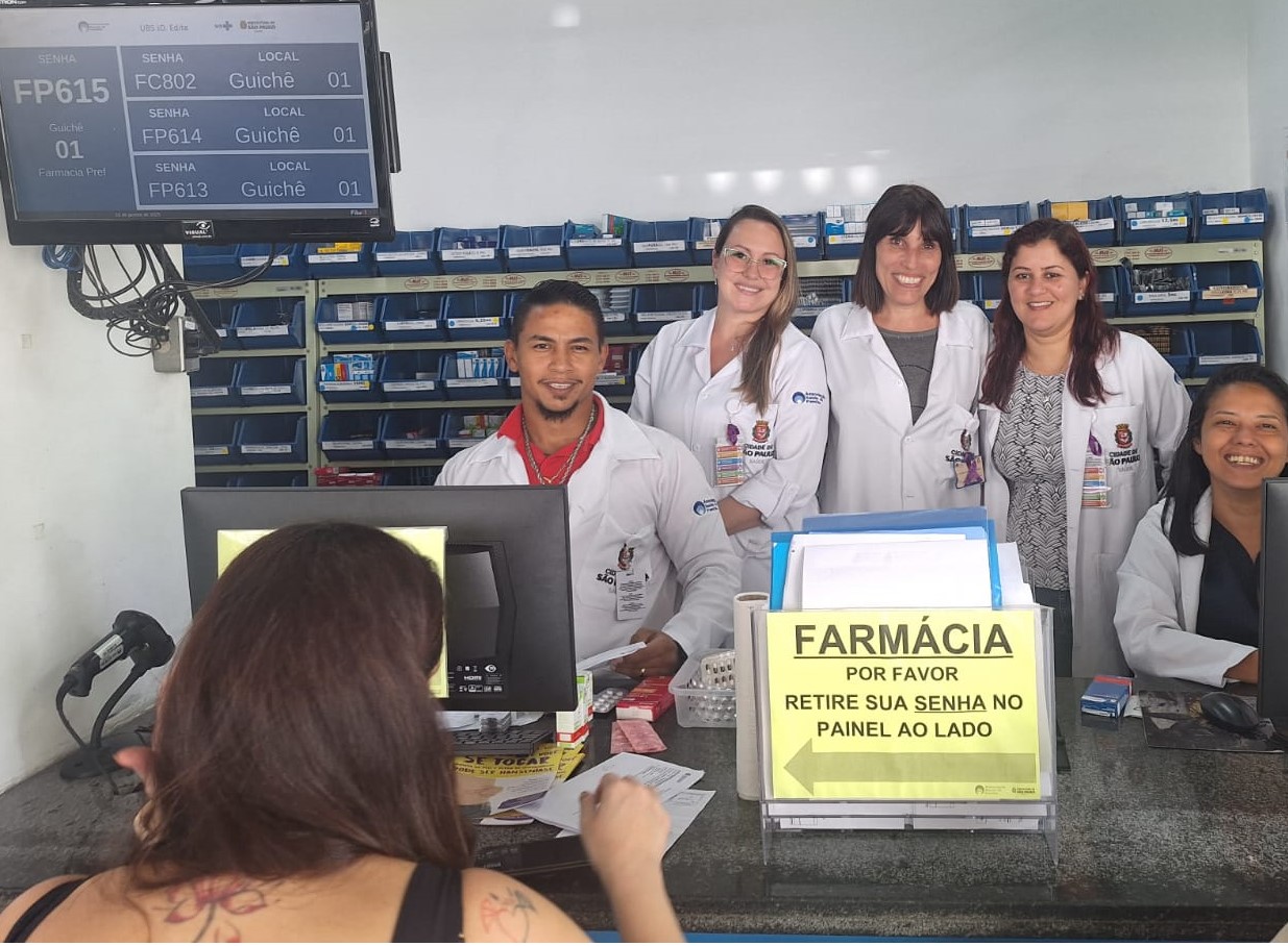 A imagem mostra uma farmácia de unidade de saúde, onde cinco profissionais, todos vestindo jalecos brancos, estão atrás de um balcão de atendimento. Eles estão sorrindo, aparentando simpatia e prontidão para atender. Há uma pessoa sendo atendida de costas para a câmera. No balcão, há um aviso com o texto: 