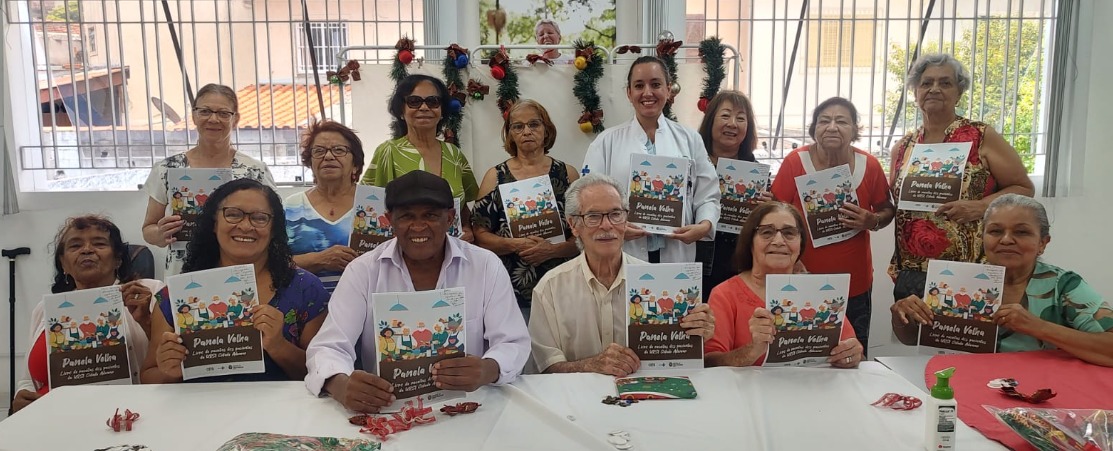 A imagem mostra um grupo de pessoas idosas reunidas em uma sala bem iluminada, provavelmente participando de uma atividade coletiva. Todos estão sentados ou em pé ao redor de uma mesa decorada com itens natalinos e seguram um livreto intitulado “Panela Velha – Com sabor e afeto”. O clima é de celebração e alegria, com sorrisos nos rostos. Ao fundo, há janelas com grades e enfeites de Natal pendurados, como bolas e festões, reforçando o tom festivo da ocasião. No centro da imagem, está uma mulher de jaleco branco, possivelmente uma profissional da saúde ou coordenadora da atividade. O cenário sugere um espaço comunitário ou de convivência, voltado para o público idoso, como um centro de saúde, centro de referência do idoso ou projeto social, onde são promovidas ações de valorização, saúde e bem-estar. O livreto parece ser parte de uma atividade educativa ou cultural, possivelmente relacionada à alimentação, memória afetiva e vínculos sociais.