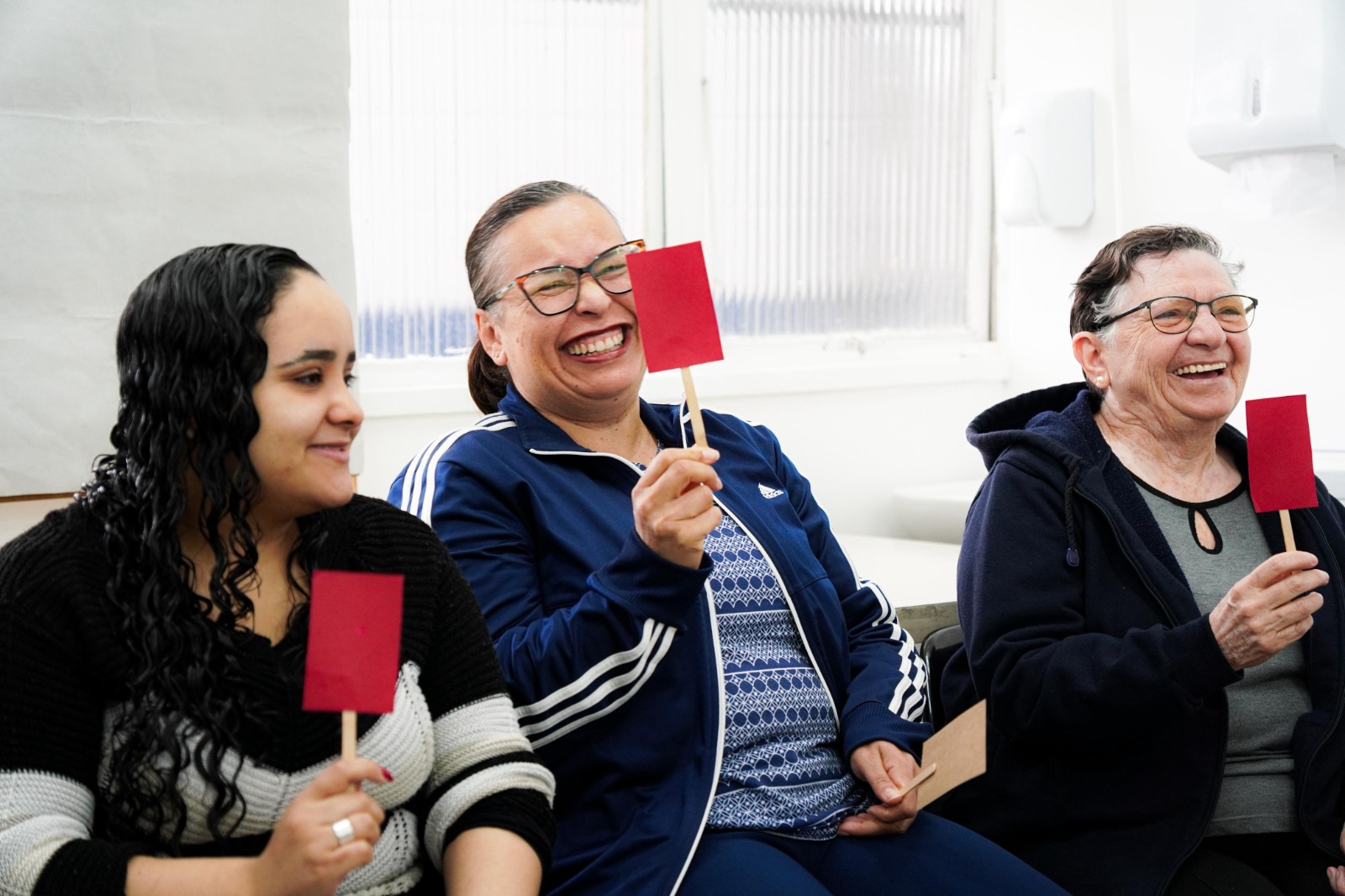 A imagem mostra três mulheres sentadas lado a lado, participando de uma atividade em grupo em um ambiente interno bem iluminado. Todas estão sorrindo e seguram pequenos cartazes vermelhos presos a palitos, levantando-os de forma descontraída. A mulher ao centro veste um agasalho azul com listras brancas e óculos; à esquerda, uma mulher de cabelos cacheados longos usa um suéter preto e cinza; à direita, uma senhora de cabelos curtos e óculos veste uma blusa cinza e um casaco azul. O clima da cena é alegre e participativo, sugerindo uma dinâmica de grupo ou oficina interativa.