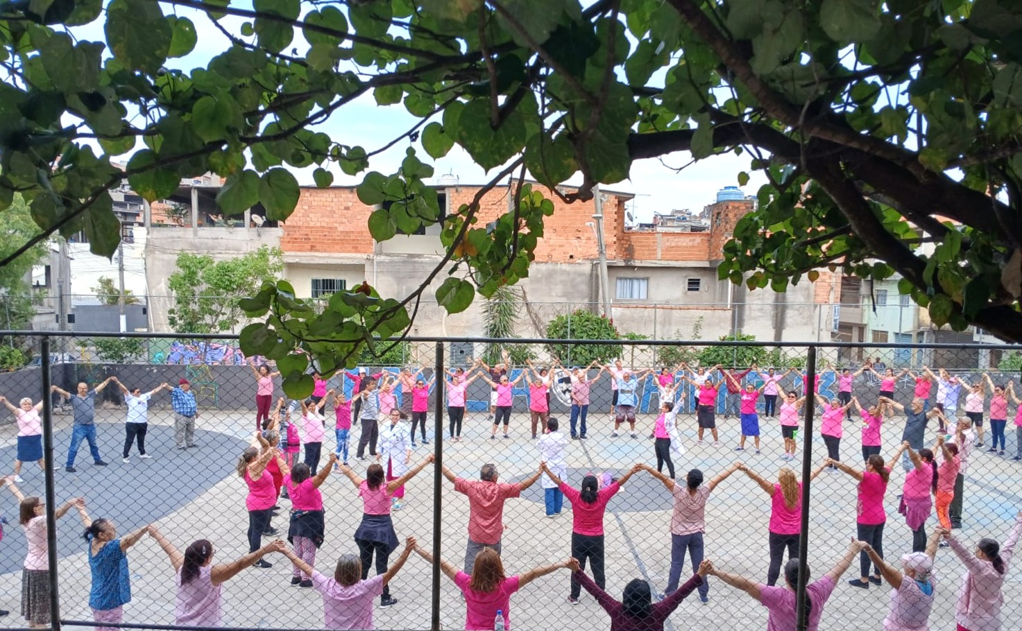 A imagem mostra um grupo grande de pessoas reunidas em uma quadra, formando um círculo amplo. Elas estão de mãos dadas e com os braços levantados, como se estivessem participando de uma atividade coletiva, possivelmente um exercício. A maioria das pessoas usa roupas em tons de rosa, o que pode indicar alguma campanha ou ação temática (como conscientização). Há participantes de diferentes idades, incluindo adultos e idosos. Ao fundo da quadra, há casas simples e construções urbanas, algumas com tijolos aparentes. Também é possível ver um muro com grafite colorido. Na parte superior da imagem, galhos e folhas de uma árvore emolduram a cena, dando um toque natural ao ambiente. A atmosfera geral transmite união, participação e atividade em grupo ao ar livre.