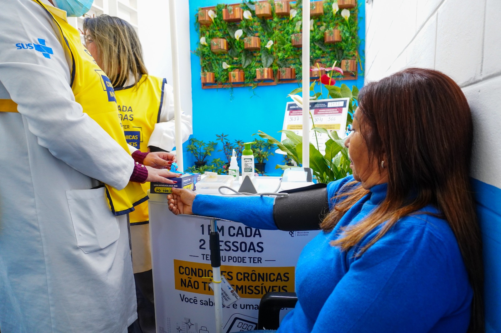 A imagem mostra o atendimento em uma Unidade Básica de Saúde durante ação de rastreamento de doenças crônicas. Uma mulher está sentada, com o braço apoiado, enquanto tem a pressão arterial aferida por um profissional de saúde. Ao redor, outros profissionais utilizam coletes de identificação da ação, e o ambiente conta com materiais de testagem e um painel informativo sobre condições crônicas não transmissíveis. Ao fundo, há um espaço organizado com plantas, reforçando um ambiente acolhedor dentro da unidade.