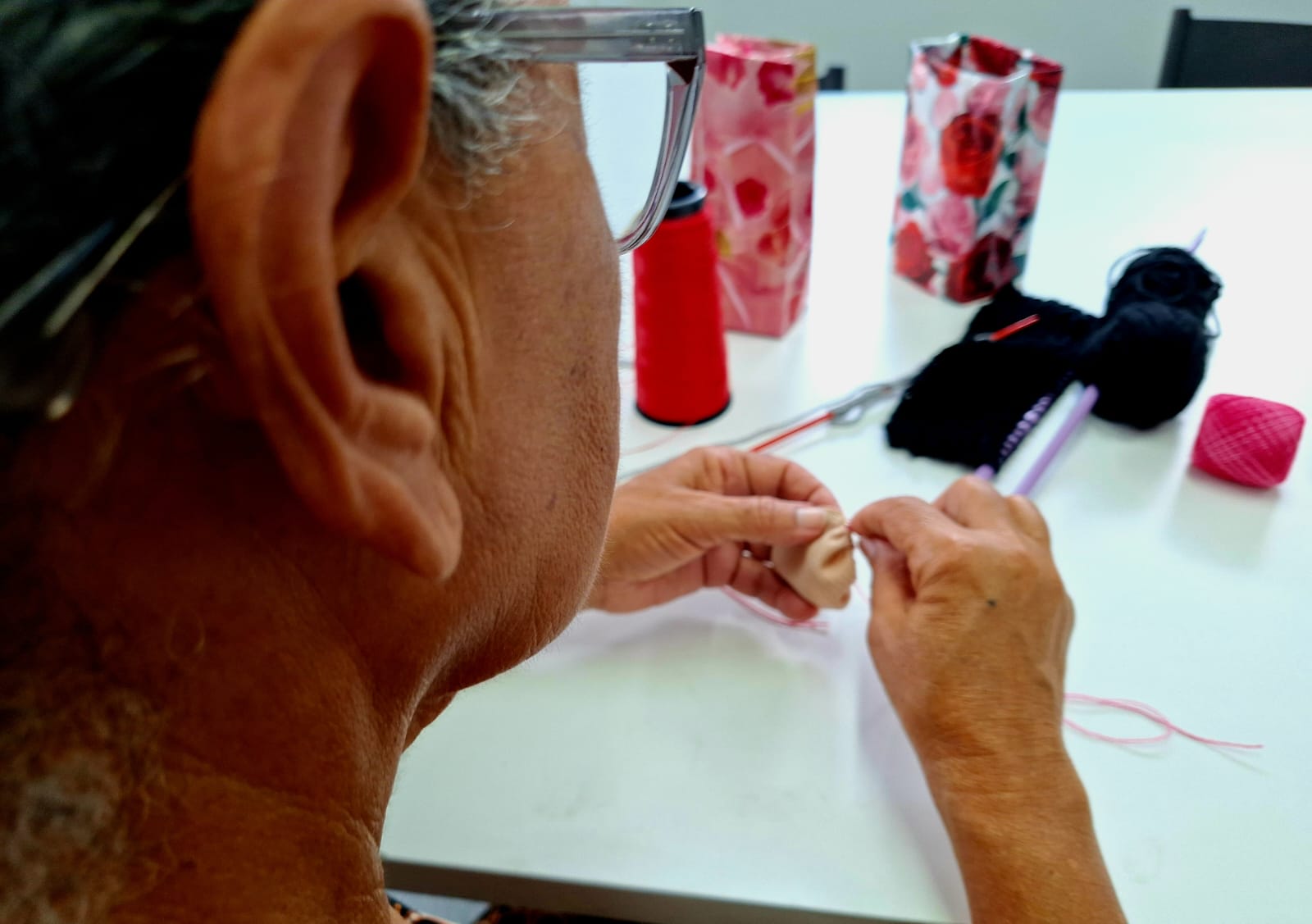 A imagem é uma fotografia colorida em formato horizontal (paisagem). Em primeiro plano, uma mulher idosa está sentada à mesa, concentrada em um trabalho de artesanato. O ambiente sugere uma sala de atividades ou escritório, com iluminação clara e direta.Seus dedos manipulam uma linha fina de cor rosa. Sobre a mesa branca, à sua frente, há materiais de costura e tricô: agulhas de plástico na cor lilás, uma agulha longa vermelha e um cone de linha vermelha vibrante à direita.
