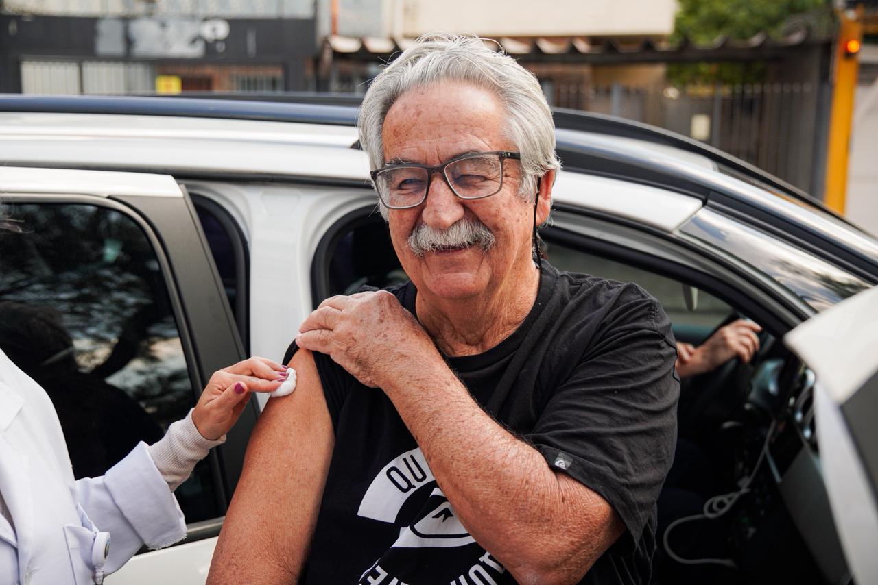 Um homem idoso, de cabelos e bigode grisalhos, usa óculos de armação escura e sorri enquanto recebe uma vacina no braço esquerdo. Vestindo uma camiseta preta de manga curta, ele segura a manga da roupa para facilitar a aplicação. Ao lado dele, aparece apenas o braço de uma profissional de saúde, com jaleco branco, pressionando um algodão no local da injeção. O atendimento acontece ao lado de um carro branco, com a porta aberta, sugerindo uma ação de vacinação em sistema drive-thru. Ao fundo, há árvores e construções urbanas desfocadas.