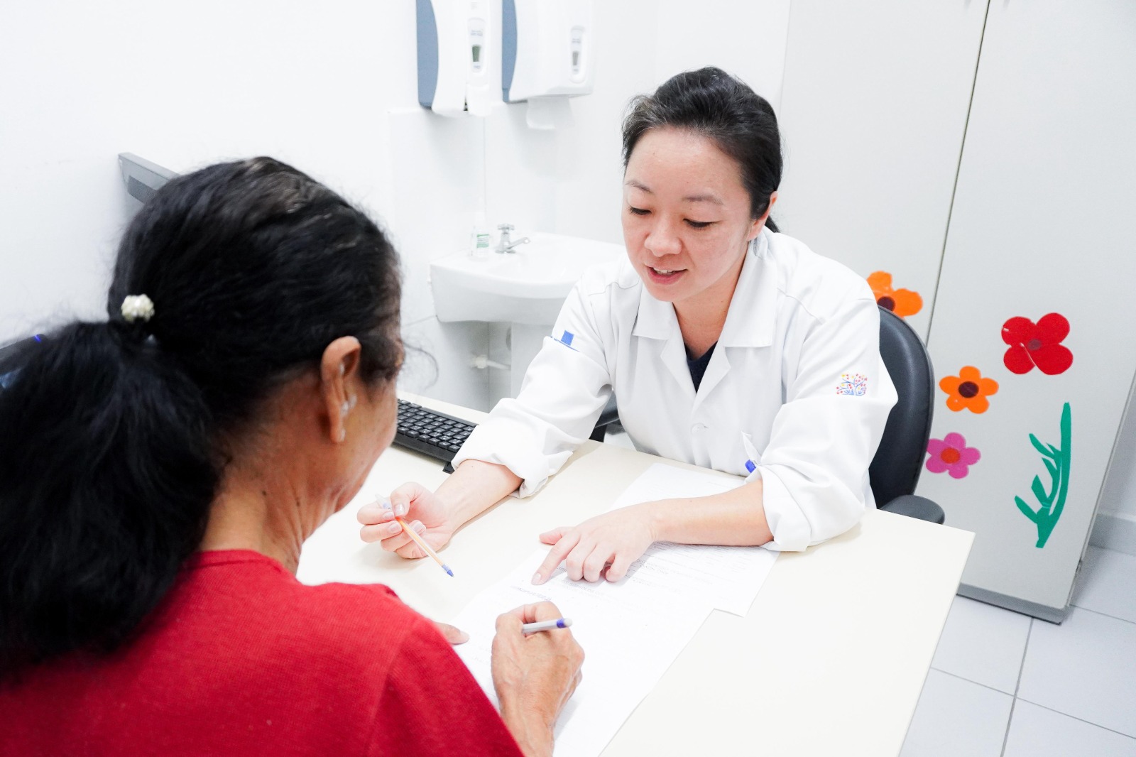 Em um consultório de saúde, uma profissional da área médica, vestindo jaleco branco, está sentada à mesa atendendo uma paciente. A profissional sorri levemente enquanto aponta para um documento sobre a mesa, ajudando a paciente, que veste blusa vermelha, a preencher o papel. O ambiente é claro e limpo, com armário ao fundo decorado com adesivos de flores coloridas.