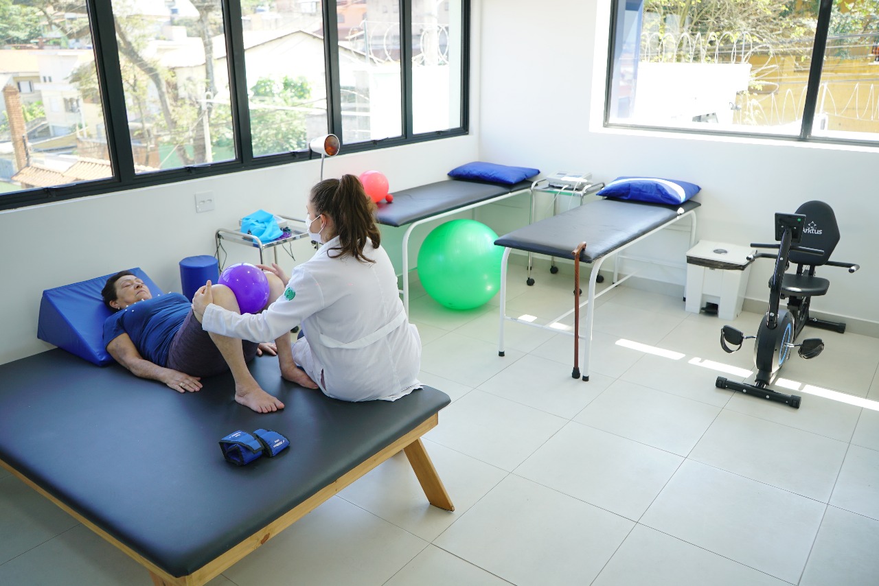 A imagem mostra uma sala clara de fisioterapia ou reabilitação, com grandes janelas que deixam entrar bastante luz natural. No centro, uma paciente idosa de pele clara e cabelos curtos e escuros está deitada sobre um colchonete preto, apoiando a cabeça em um triângulo azul de espuma. Ela veste camiseta azul e shorts roxos.  À frente dela, uma profissional de saúde, de pele clara e cabelos castanhos presos em rabo de cavalo, usa jaleco branco e máscara, conduzindo um exercício. A fisioterapeuta segura uma bola roxa entre os joelhos da paciente, orientando o movimento de fortalecimento.  No fundo da sala, há duas macas com travesseiros azuis, uma bola verde de pilates, um andador, um bastão de caminhada encostado, e uma bicicleta ergométrica. O espaço é limpo, organizado e voltado à prática de exercícios de reabilitação.