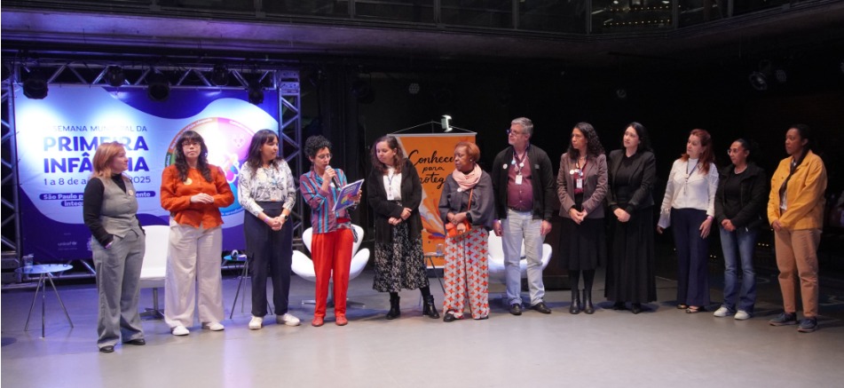 A imagem mostra um grupo de 12 pessoas enfileiradas em um palco, aparentemente participando de um evento oficial. No fundo, há um grande painel iluminado com a inscrição “Semana Municipal da Primeira Infância – 1 a 8 de agosto de 2025”, com o brasão da cidade de São Paulo e menção ao “Plano Municipal da Primeira Infância”.  À direita do palco, há um banner laranja com a frase “Conhecer para proteger” e a imagem estilizada de uma criança.  O grupo é diverso, formado por mulheres e um homem, com trajes formais ou casuais. Uma das mulheres, ao centro, está com um microfone e lendo um papel, indicando que é a pessoa que fala no momento. O clima é de solenidade ou abertura de um evento institucional relacionado à infância, com foco em políticas públicas, proteção e desenvolvimento infantil.