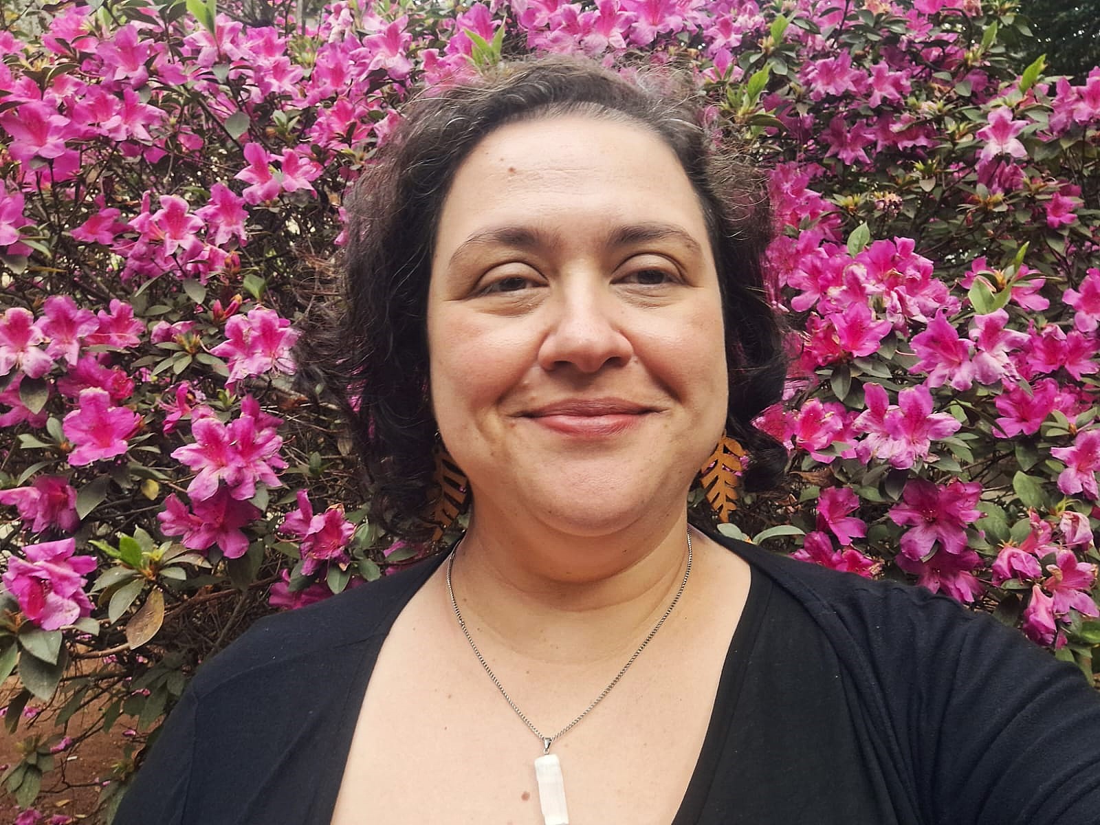 Foto em close (do peito para cima) de Camila, uma pessoa adulta em ambiente externo. Ela tem pele clara, cabelo curto, escuro e ondulado, com alguns fios soltos. O rosto é arredondado, com sobrancelhas escuras, olhos castanhos, e sorriso discreto de lábios fechados, transmitindo expressão tranquila e simpática. Veste blusa/preto com decote em “U” e usa um colar de corrente fina prateada com pingente retangular branco translúcido (parece pedra/pau-brasil lapidado). Nos ouvidos, brincos laranja em formato de folha, com recortes que lembram nervuras.  Ao fundo, ocupando toda a imagem, há um arbusto denso de flores rosa-vibrante (parecem azaleias), com folhas verdes escuras. A luz é natural e difusa, sem sombras duras, sugerindo dia nublado ou sombra de árvores.