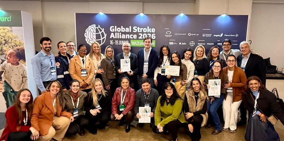 A foto mostra um grande grupo de pessoas reunidas em um evento, posando de forma descontraída e comemorativa diante de um painel institucional. Ao fundo, há um banner com a inscrição “Global Stroke Alliance 2026”, além das datas do evento e diversos logotipos de organizações apoiadoras. O grupo é composto por homens e mulheres de diferentes idades, muitos usando crachás de identificação pendurados no pescoço, indicando participação em um congresso ou encontro profissional. As pessoas estão distribuídas em duas fileiras: algumas em pé e outras agachadas na parte da frente. Vários participantes levantam os braços e acenam para a câmera, transmitindo entusiasmo, integração e celebração. Algumas pessoas seguram certificados ou placas de reconhecimento, sugerindo uma premiação, apresentação de projetos ou destaque em atividades do evento. O ambiente é interno, com iluminação clara e profissional. As expressões são predominantemente sorridentes e alegres, reforçando o clima de confraternização e conquista coletiva. No canto inferior direito, é possível ver parte de um celular fotografando a mesma cena, indicando que o momento estava sendo registrado por diferentes participantes.