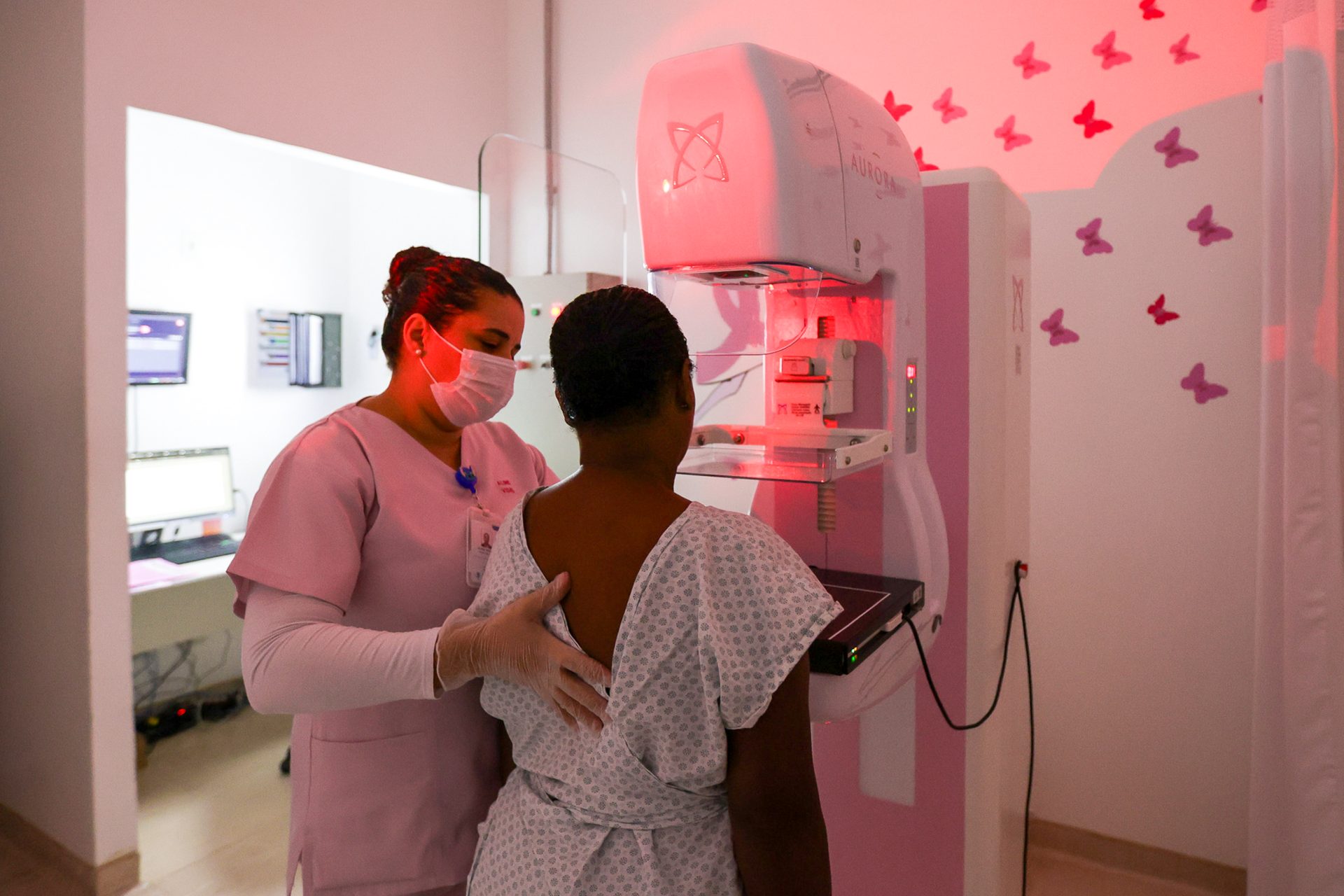 Uma profissional de saúde vestida com uniforme rosa e máscara facial posiciona uma paciente para realizar um exame de mamografia. A paciente, de costas para a câmera, veste um avental hospitalar aberto na parte de trás e está em frente ao aparelho de mamografia, que emite uma luz avermelhada. O ambiente é limpo e bem iluminado, com paredes brancas decoradas com adesivos de borboletas cor-de-rosa e lilás. Ao fundo, há uma sala anexa com computadores e monitores ligados, visíveis através de uma janela de vidro. A profissional apoia gentilmente as costas da paciente, orientando-a durante o procedimento.
