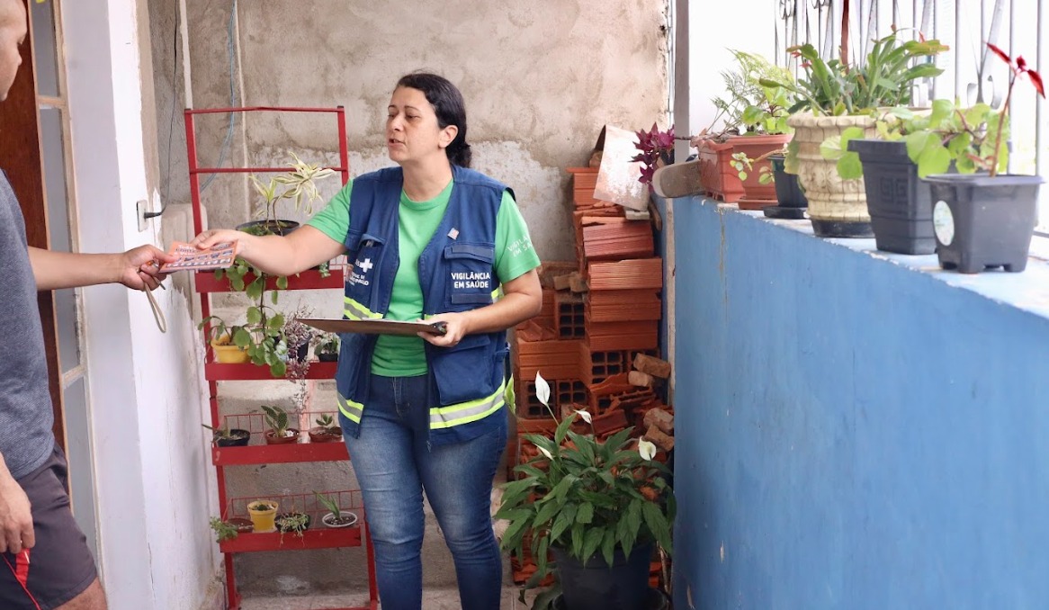 A imagem mostra uma agente de saúde em visita domiciliar, conversando com um morador em uma área externa de uma casa. Ela veste camiseta verde e colete azul com identificação de “Vigilância em Saúde”, segura uma prancheta e entrega um material informativo ao homem, que aparece parcialmente à esquerda. O ambiente é simples, com paredes sem acabamento completo e um pequeno espaço com plantas em vasos — alguns organizados em uma estante vermelha e outros sobre um muro azul. Há também tijolos empilhados ao fundo, sugerindo obra ou adaptação do espaço.