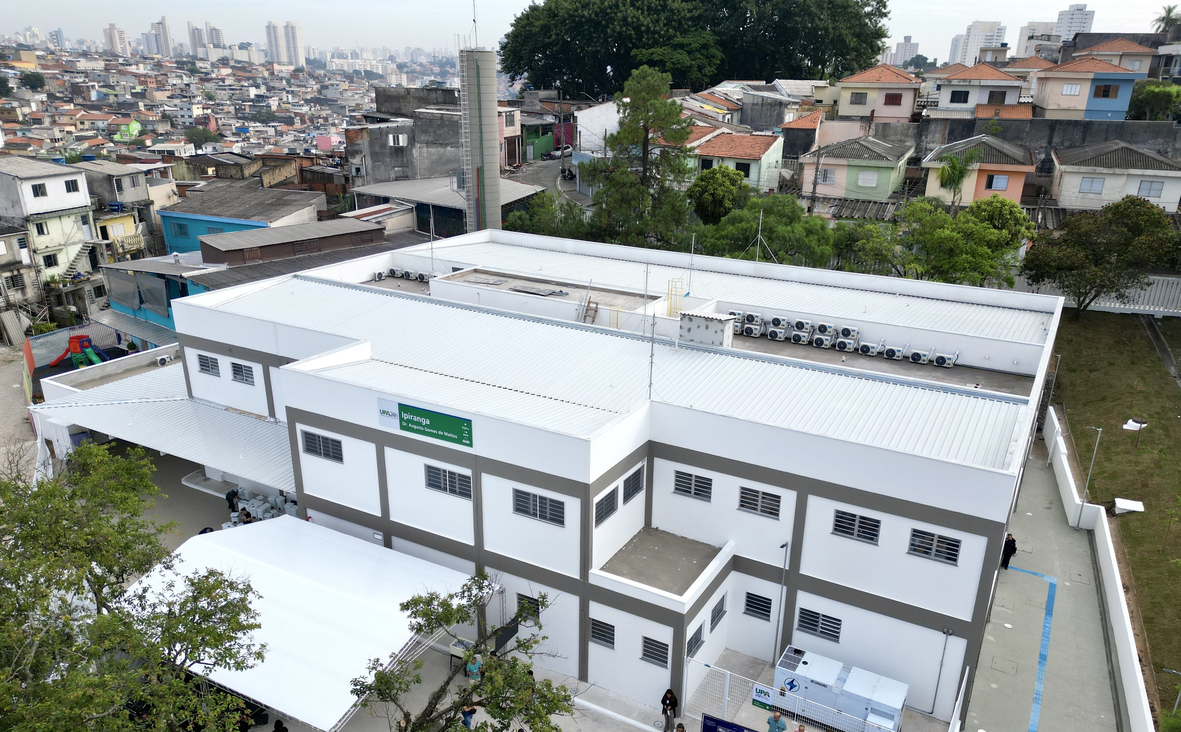 A imagem mostra uma unidade de saúde de médio porte, vista de cima, em um bairro urbano. O prédio é novo, com pintura branca e detalhes em cinza, telhado metálico e diversos aparelhos de ar-condicionado instalados na parte superior. Na fachada lateral, há uma placa verde com o nome UPA Ipiranga – Unidade de Pronto Atendimento 24h. Em frente ao prédio, foi montada uma estrutura com cobertura branca, provavelmente para abrigar visitantes ou a realização de um evento inaugural. O entorno é composto por casas simples e coloridas, dispostas em ruas estreitas e irregulares, com uma área de maior adensamento urbano ao fundo, onde se veem prédios residenciais mais altos. A cena sugere a integração da unidade de saúde com a comunidade local, em uma região de grande densidade populacional.