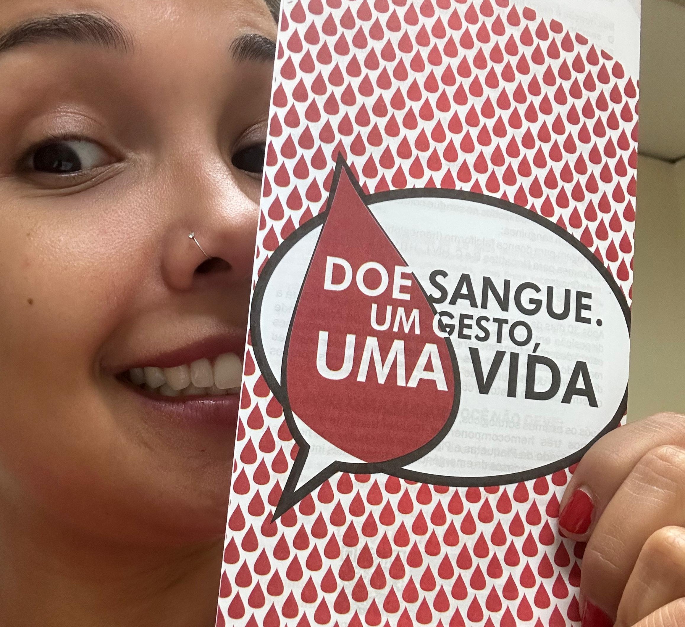 A imagem mostra uma mulher sorridente segurando um panfleto sobre doação de sangue. O panfleto tem um fundo repleto de gotas vermelhas, simbolizando sangue, e uma grande gota com um balão de fala contendo a mensagem: