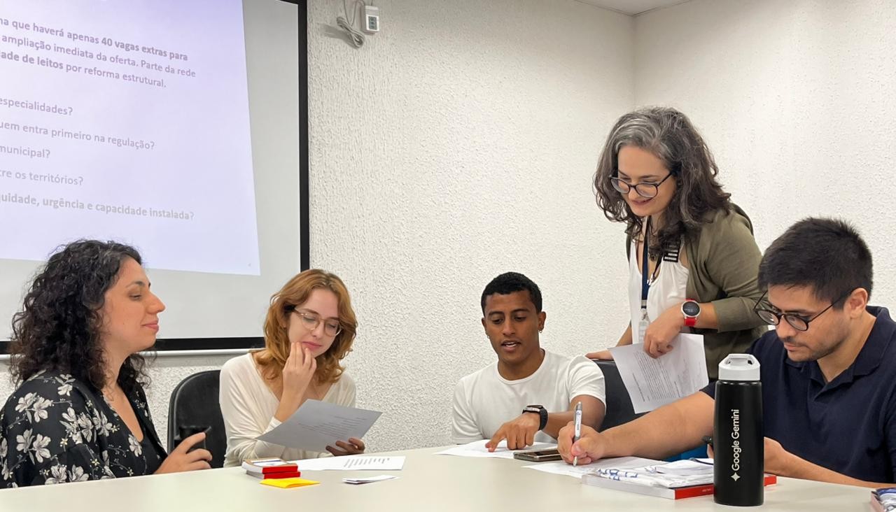A imagem mostra um grupo de cinco pessoas reunidas em uma sala, aparentemente em um ambiente de trabalho ou estudo, como uma reunião ou atividade em grupo. Elas estão sentadas ao redor de uma mesa, com papéis e materiais espalhados à frente. Quatro pessoas estão sentadas, enquanto uma mulher está em pé, levemente inclinada, observando e interagindo com o grupo - possivelmente orientando ou participando ativamente da discussão. No fundo da sala há uma parede clara e uma tela de projeção com um slide exibindo texto, o que reforça a ideia de que se trata de uma apresentação ou dinâmica de aprendizado. As expressões das pessoas indicam concentração e colaboração. Sobre a mesa, há itens como papéis, canetas e celular. 