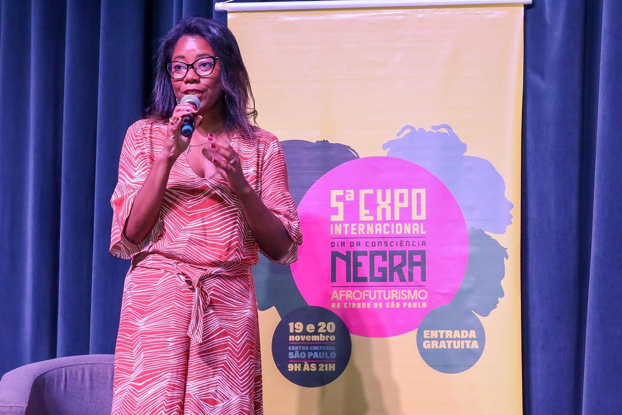 Doutora Lígia Mascarenhas, uma mulher negra, está em pé falando ao microfone em um palco. Ela usa óculos e um macacão laranja com estampa geométrica branca. Ao fundo, há um painel amarelo com o texto “5ª Expo Internacional Dia da Consciência Negra – Afrofuturismo na Cidade de São Paulo”, acompanhado de silhuetas de perfis humanos em azul. O painel também exibe informações do evento: “19 e 20 de novembro, Centro Cultural São Paulo, 9h às 21h” e “Entrada gratuita”. A cortina do palco é azul escura.