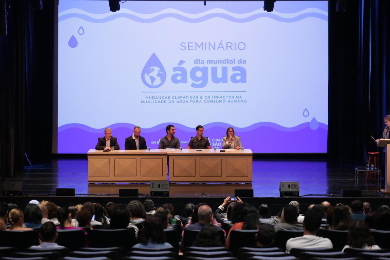 No fundo um painel na cor azul destaca o título do evento: Seminário Dia Mundial da Água - Mudanças Climáticas e os Impactos na Qualidade da Água para Consumo Humano. Na mesa de abertura, da esquerda para a direita, o diretor de Meio Ambiente do Centro de Vigilância Sanitária do Estado de São Paulo, representante da Agência Nacional da Água (ANA), da Coordenadoria de Vigilância em Saúde (Covisa), do Ministério da SaúdeI(MS) e da Diretora da Divisão de Saúde Ambiental (DVISAM) da Secretaria Municipal da Saúde (SMS)