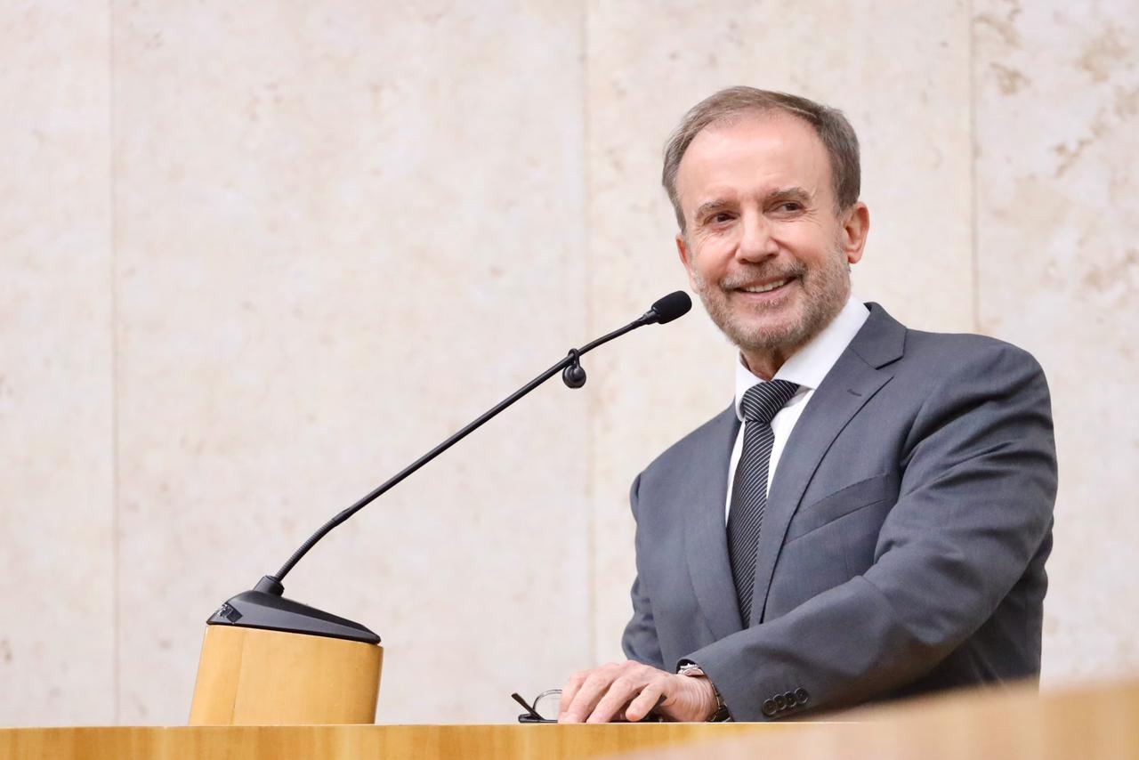 Secretario  Municipal da Saúde, Luiz Carlos Zamarco, discursa no púlpito da Câmara Municipal de São Paulo. Ele esta vestindo um terno cinza e está sorrindo e segundo um óculos.   