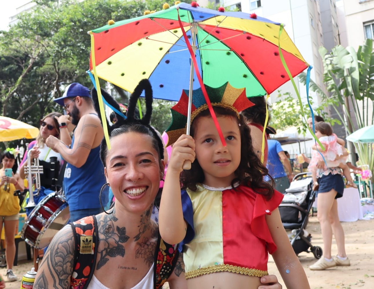 A imagem mostra uma cena alegre ao ar livre, de um bloco infantil de Carnaval em uma praça. No centro, há uma mulher sorridente com tatuagens nos braços, usando uma tiara preta com orelhinhas. Ao lado dela está uma criança vestida com uma fantasia colorida, segurando um guarda-chuva pequeno e com cores vibrantes como vermelho, azul, verde e amarelo. A criança também usa uma espécie de adereço na cabeça que combina com o clima festivo. Ao fundo, é possível ver várias pessoas, incluindo crianças e adultos, algumas também fantasiadas. Há um músico com um tambor, outras pessoas segurando instrumentos e um carrinho de bebê.