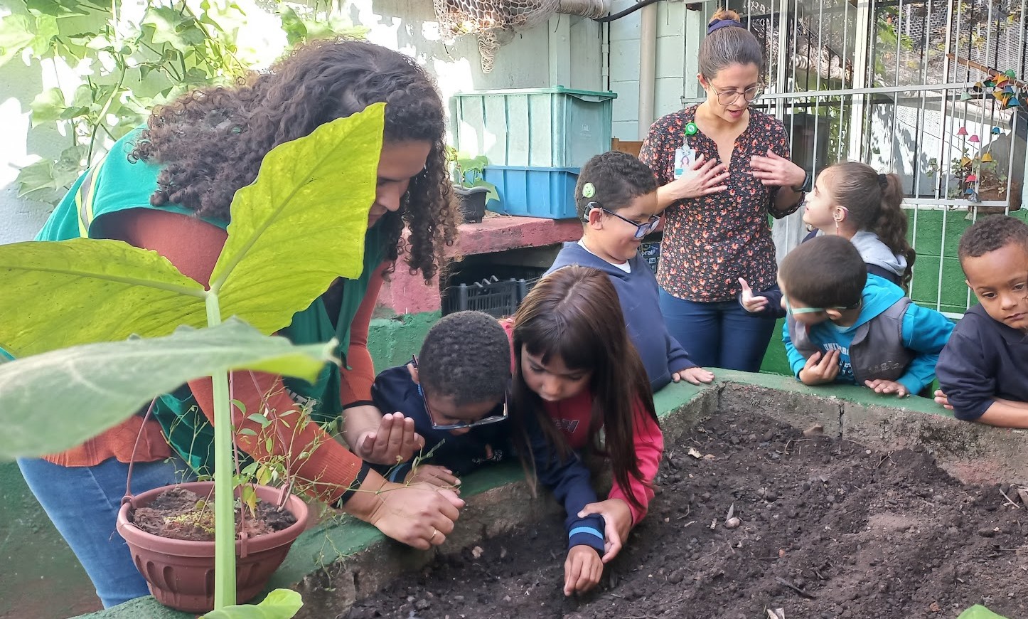 A imagem mostra um grupo de crianças participando de uma atividade em uma horta, acompanhadas por duas profissionais. Em primeiro plano, algumas crianças estão curvadas sobre o canteiro de terra, observando e mexendo no solo com atenção, como se estivessem plantando ou procurando sementes. À esquerda, uma profissional de colete verde auxilia de perto duas crianças, incentivando a participação delas. Ao fundo, outra profissional, de óculos e camisa estampada, conversa com as crianças, que a escutam atentamente. O ambiente tem plantas, vasos e caixas organizadas, transmitindo a ideia de um espaço educativo e acolhedor. A cena transmite aprendizado coletivo, cuidado com a natureza e inclusão, mostrando a horta como recurso pedagógico para despertar a consciência ambiental e o trabalho em grupo.