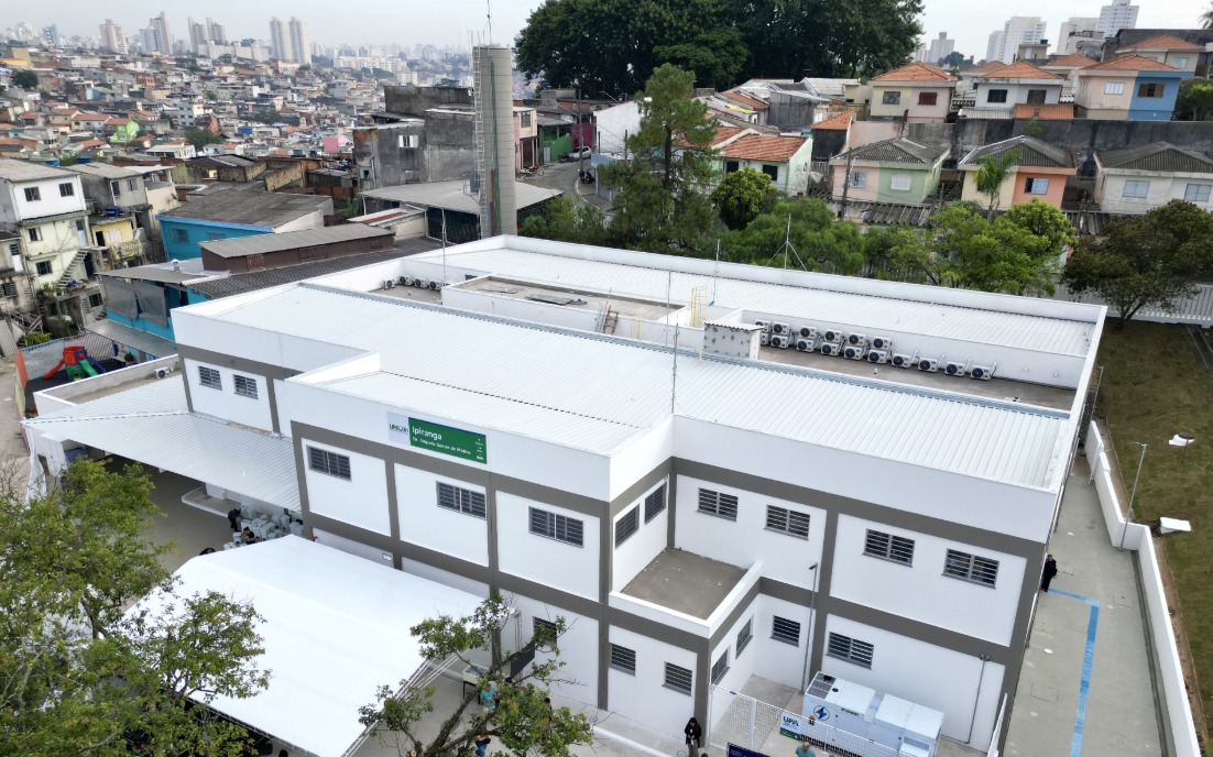  A imagem mostra uma unidade de saúde de médio porte, vista de cima, em um bairro urbano. O prédio é novo, com pintura branca e detalhes em cinza, telhado metálico e diversos aparelhos de ar-condicionado instalados na parte superior. &nbsp;Na fachada lateral, há uma placa verde com o nome UPA Ipiranga – Unidade de Pronto Atendimento 24h. Em frente ao prédio, foi montada uma estrutura com cobertura branca, provavelmente para abrigar visitantes ou a realização de um evento inaugural. &nbsp;O entorno é composto por casas simples e coloridas, dispostas em ruas estreitas e irregulares, com uma área de maior adensamento urbano ao fundo, onde se veem prédios residenciais mais altos. &nbsp;A cena sugere a integração da unidade de saúde com a comunidade local, em uma região de grande densidade populacional.