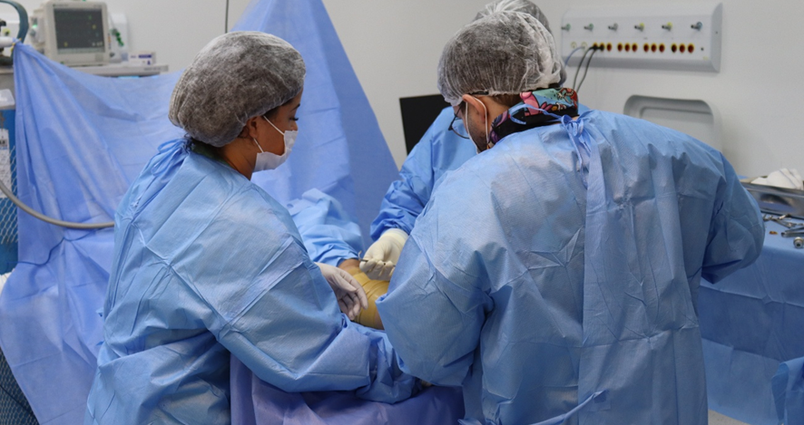 A imagem mostra uma equipe médica realizando um procedimento cirúrgico em um ambiente hospitalar. Três profissionais da saúde, vestidos com roupas cirúrgicas esterilizadas de cor azul e toucas descartáveis, estão concentrados na realização da cirurgia. Eles utilizam luvas e máscaras, seguindo os protocolos de segurança e higiene. Ao fundo, é possível ver equipamentos médicos, como monitores e uma mesa com instrumentos cirúrgicos. A cena representa um momento de cuidado e precisão, evidenciando o trabalho conjunto da equipe de saúde no centro cirúrgico