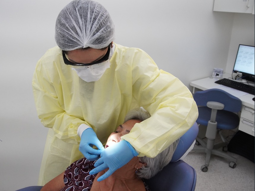 A imagem mostra uma profissional de saúde realizando atendimento odontológico em uma paciente idosa. A profissional usa equipamentos de proteção individual, incluindo touca, avental amarelo, máscara, óculos de proteção e luvas azuis. A paciente está sentada na cadeira odontológica, com a cabeça apoiada, enquanto a profissional examina sua boca. No fundo, há uma mesa com computador, teclado e cadeira, compondo um consultório odontológico limpo e bem iluminado.