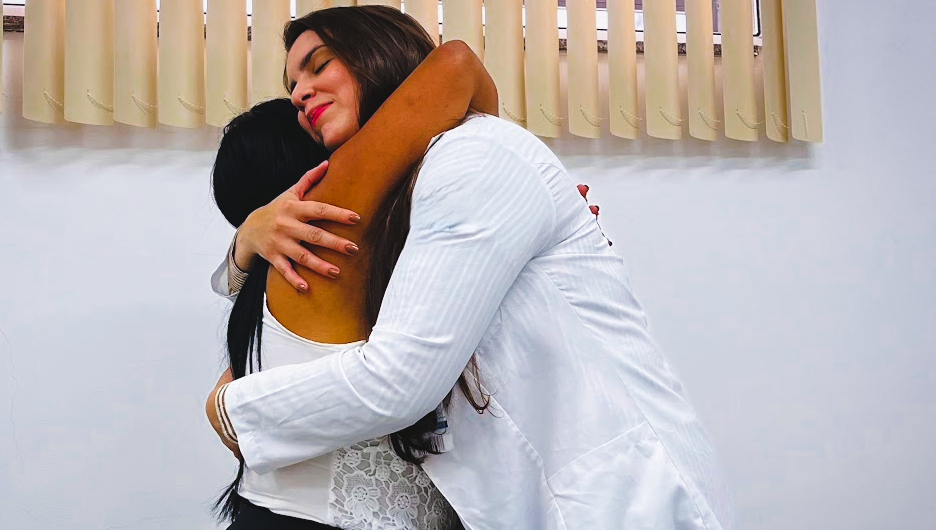 Duas mulheres se abraçam em um ambiente de saúde. Uma delas usa jaleco branco e a outra está de costas para a câmera. A cena transmite acolhimento, afeto e vínculo entre profissional de saúde e paciente.